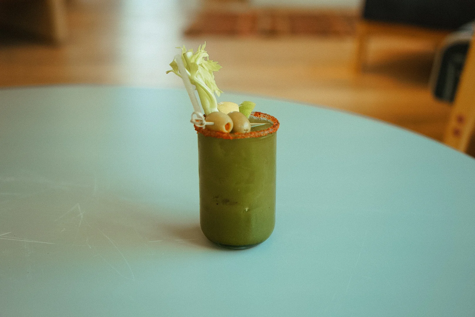 A cocktail glass filled with a green drink garnished with celery and olives, sitting on a light-colored table.