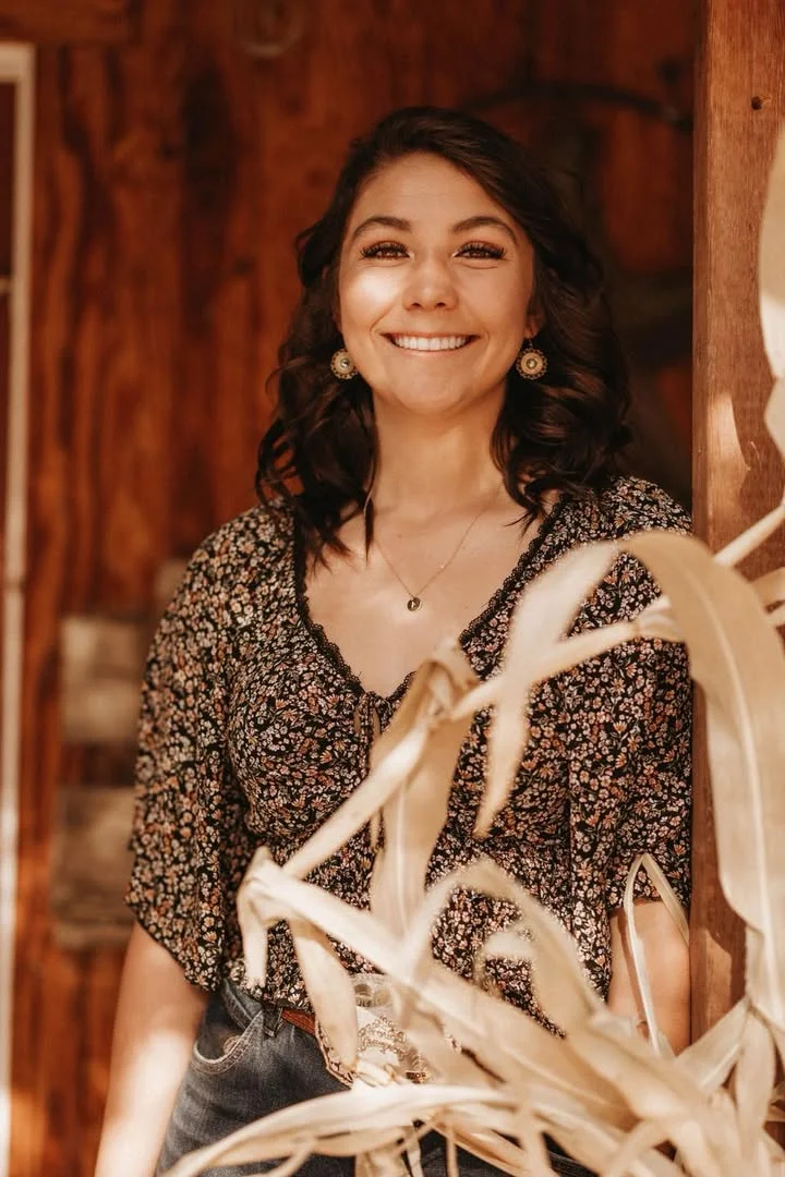 A cheerful person standing beside a wooden structure, framed by dried plants.