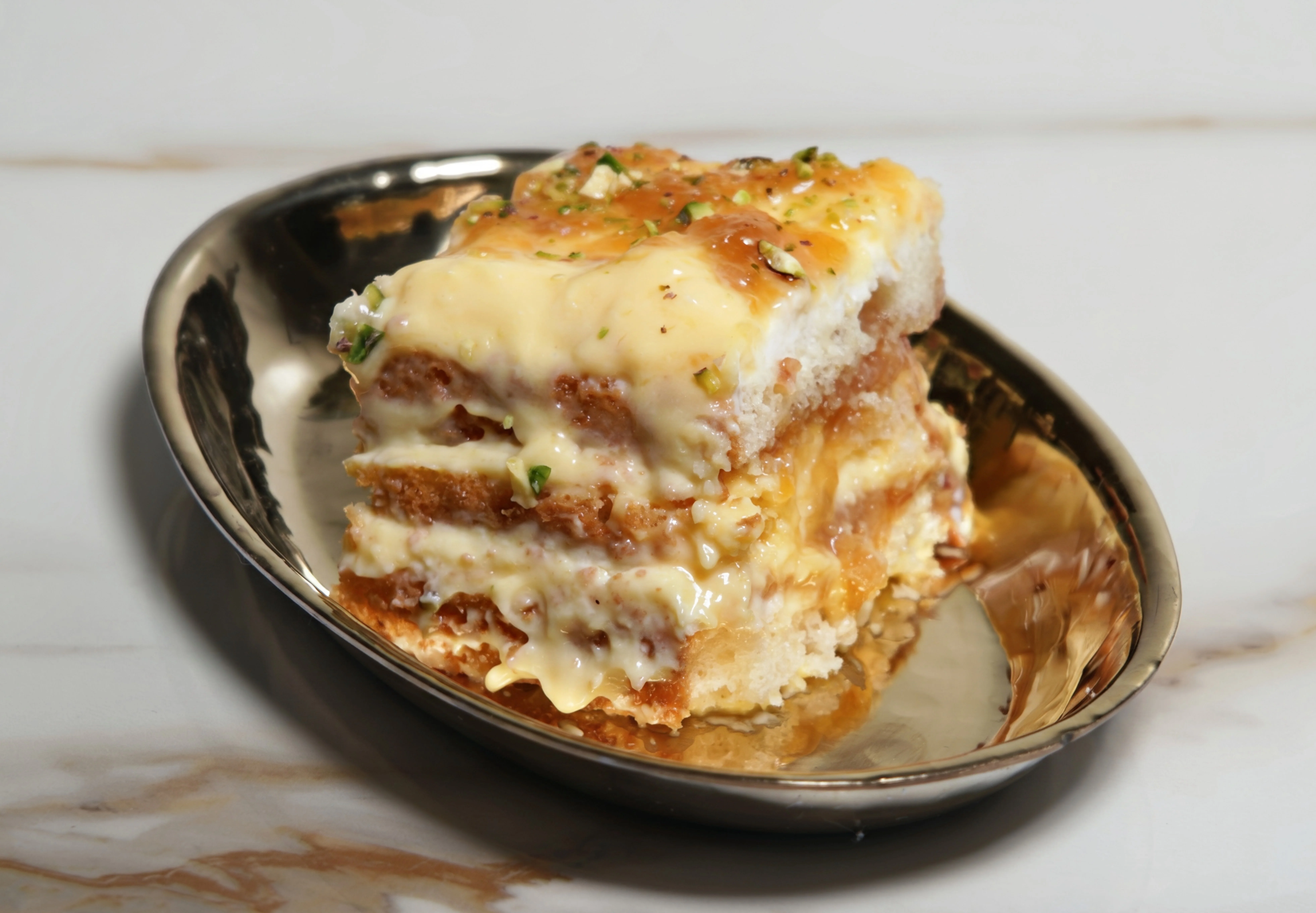 A slice of creamy dessert stacked with layers, showcasing a rich filling and a sprinkle of pistachios on top. It is served on a shiny metal plate.