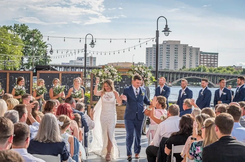 A joyful wedding ceremony taking place by a waterfront, with the bride and groom walking down the aisle amidst cheering guests and floral decorations.