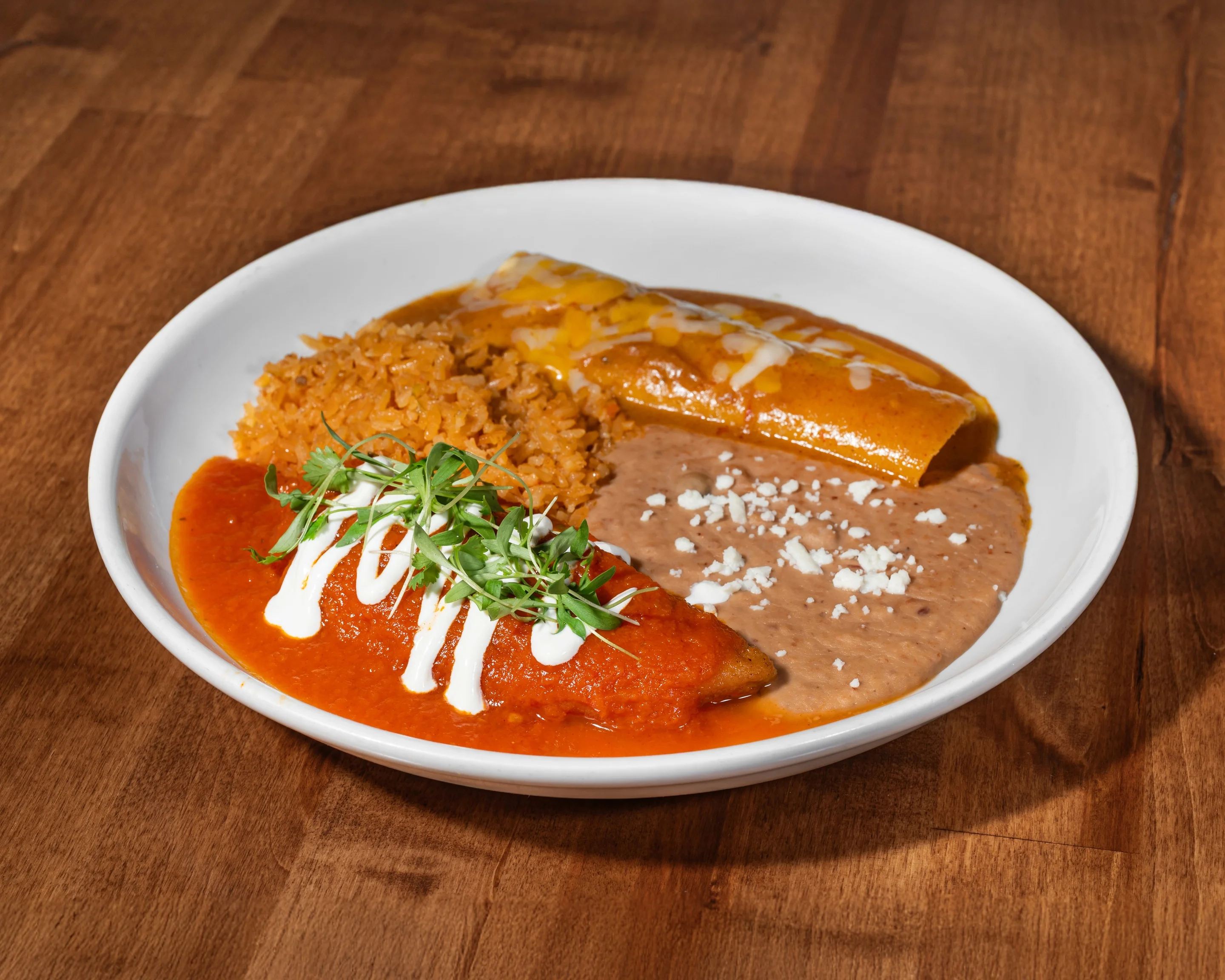 A plate of enchiladas topped with red sauce and cheese, served with refried beans and orange rice, garnished with sour cream and herbs.
