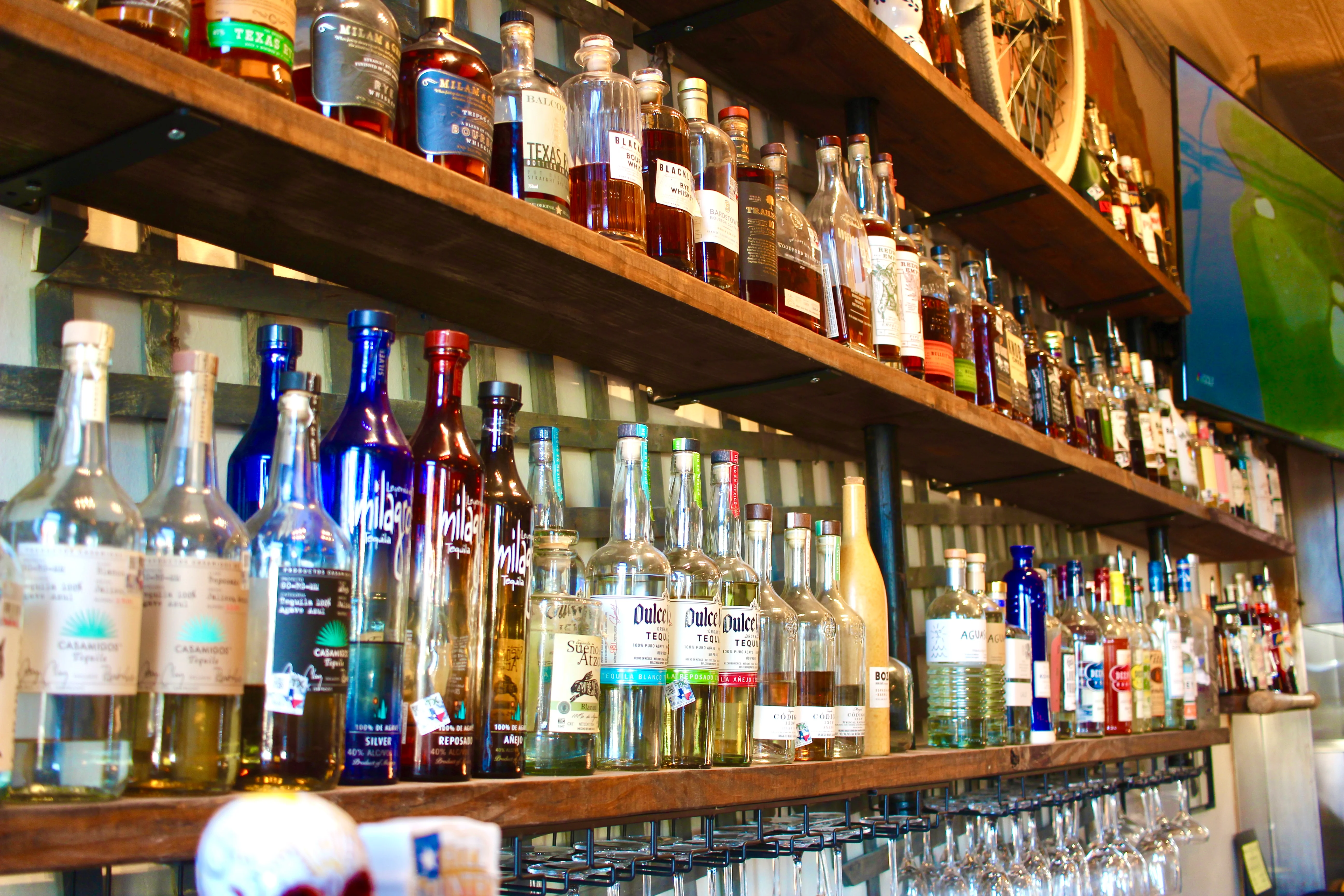 A well-stocked bar shelf displaying a variety of liquor bottles, including tequila, whiskey, and vodka, with glasses hanging below.