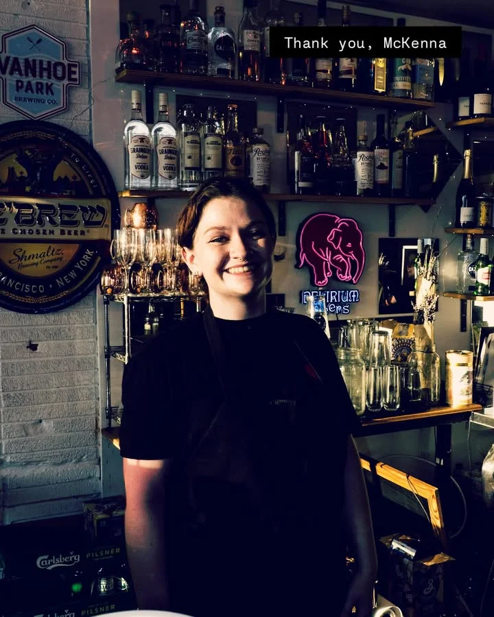 A smiling bartender stands in front of a well-stocked bar with various bottles and glasses visible.