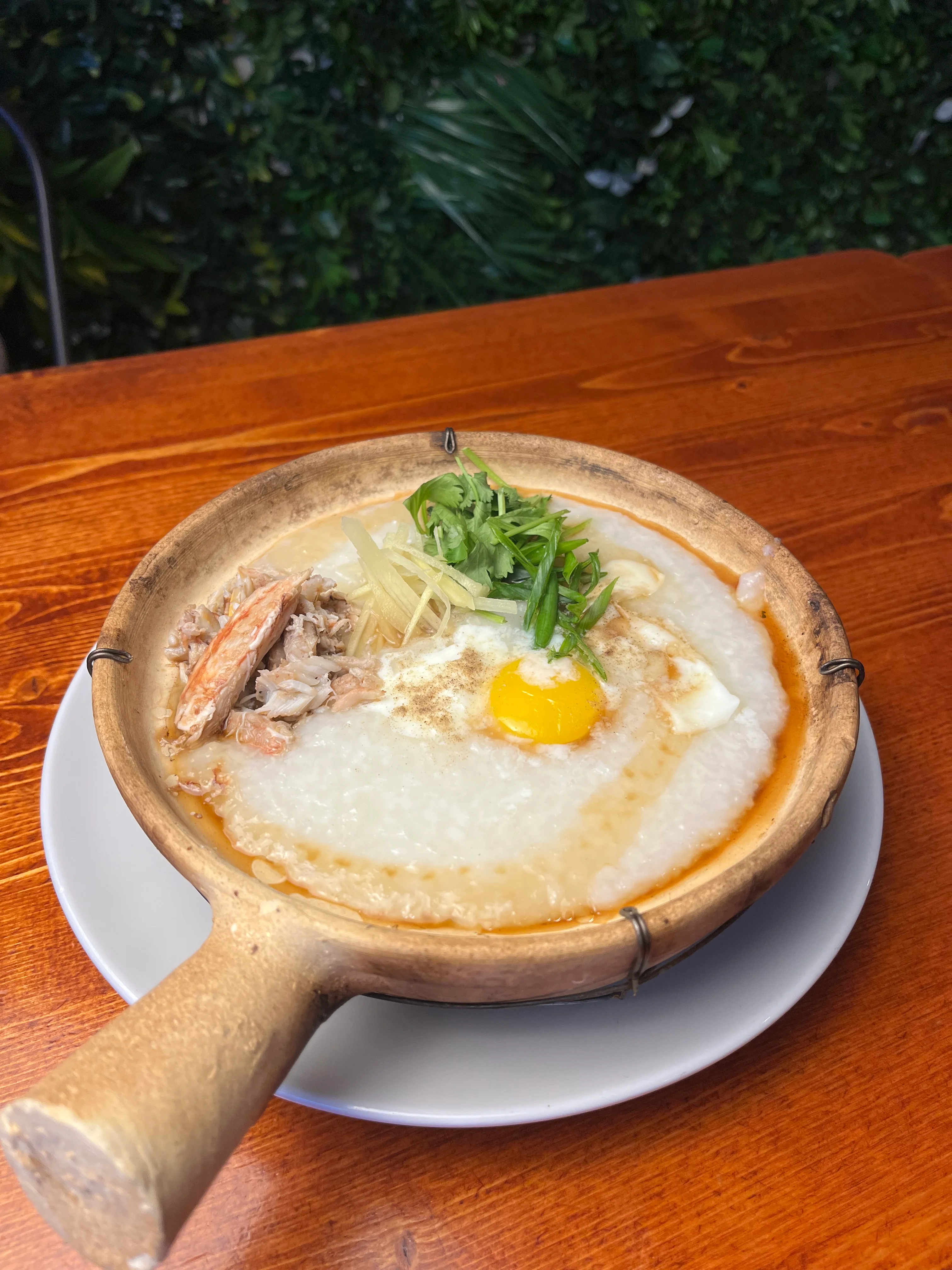A clay pot filled with rice porridge, topped with shredded meat, a raw egg, and garnished with ginger and green onions.