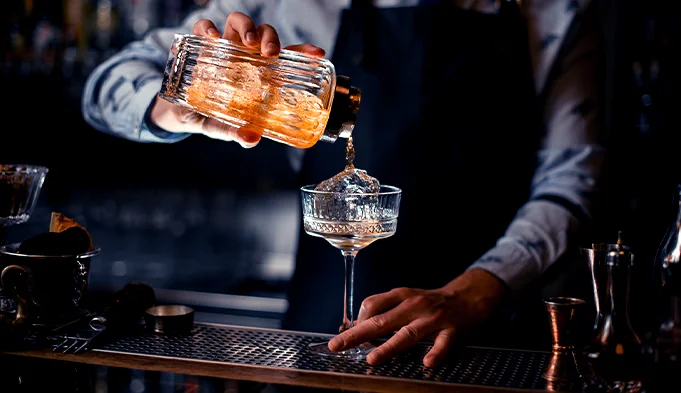 A bartender preparing cocktails at a bar with various bottles in the background.