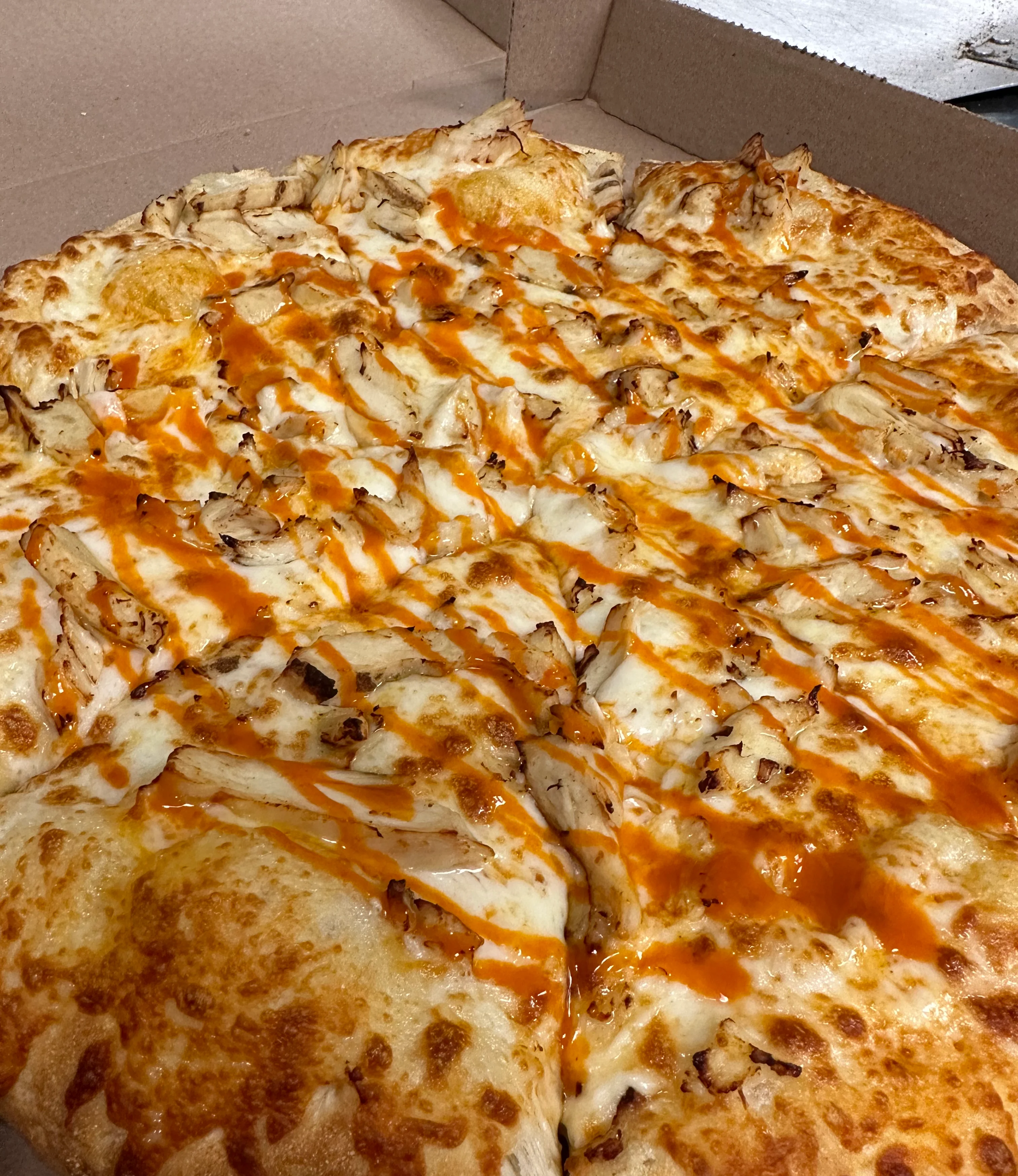 A round pizza with a brown crust, topped with cheese and drizzled with barbecue sauce. The pizza is presented in a cardboard box.