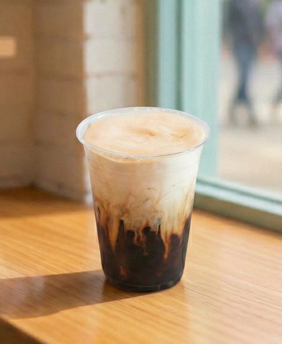 An iced coffee with cream on a wooden counter in a bright coffee shop with a blurred springtime cherry blossom background.