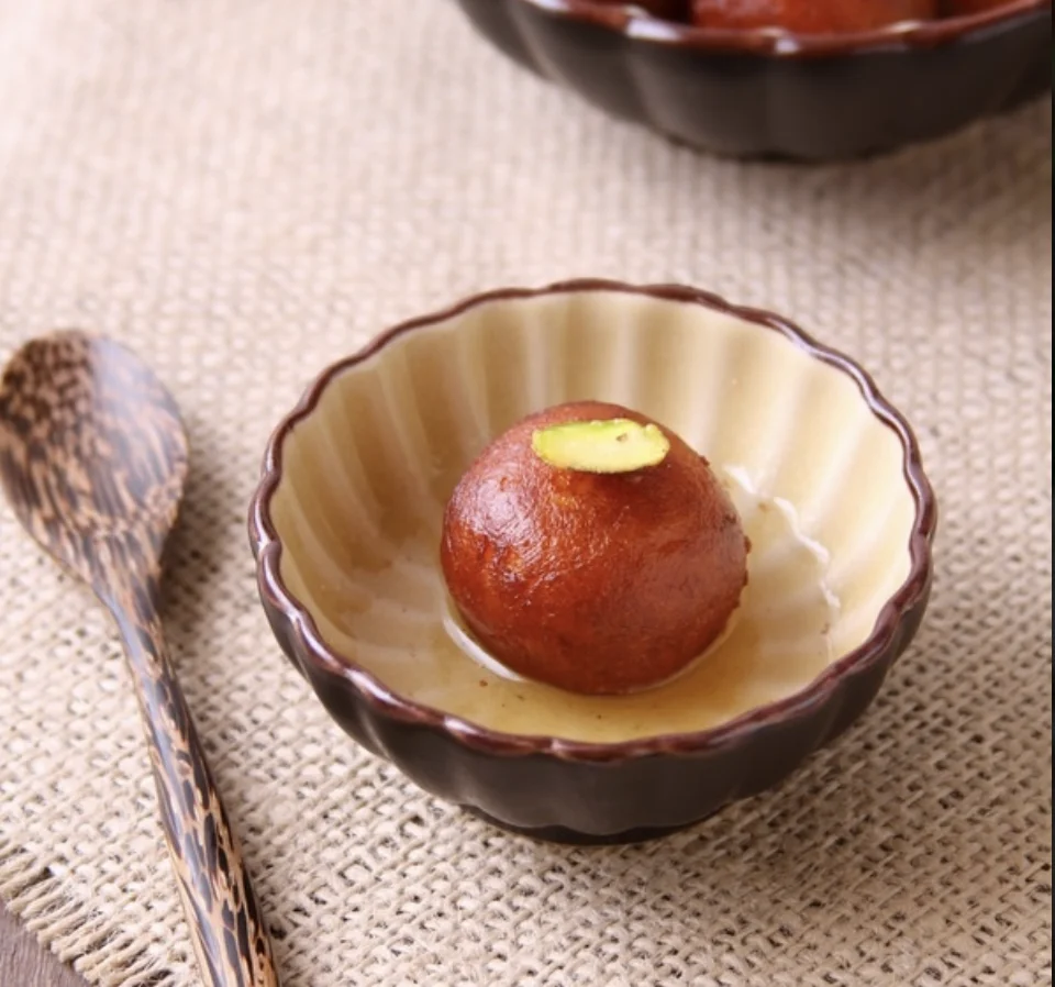 A single round Indian sweet dumpling soaked in syrup, served in a small dish with a slice of pistachio on top.