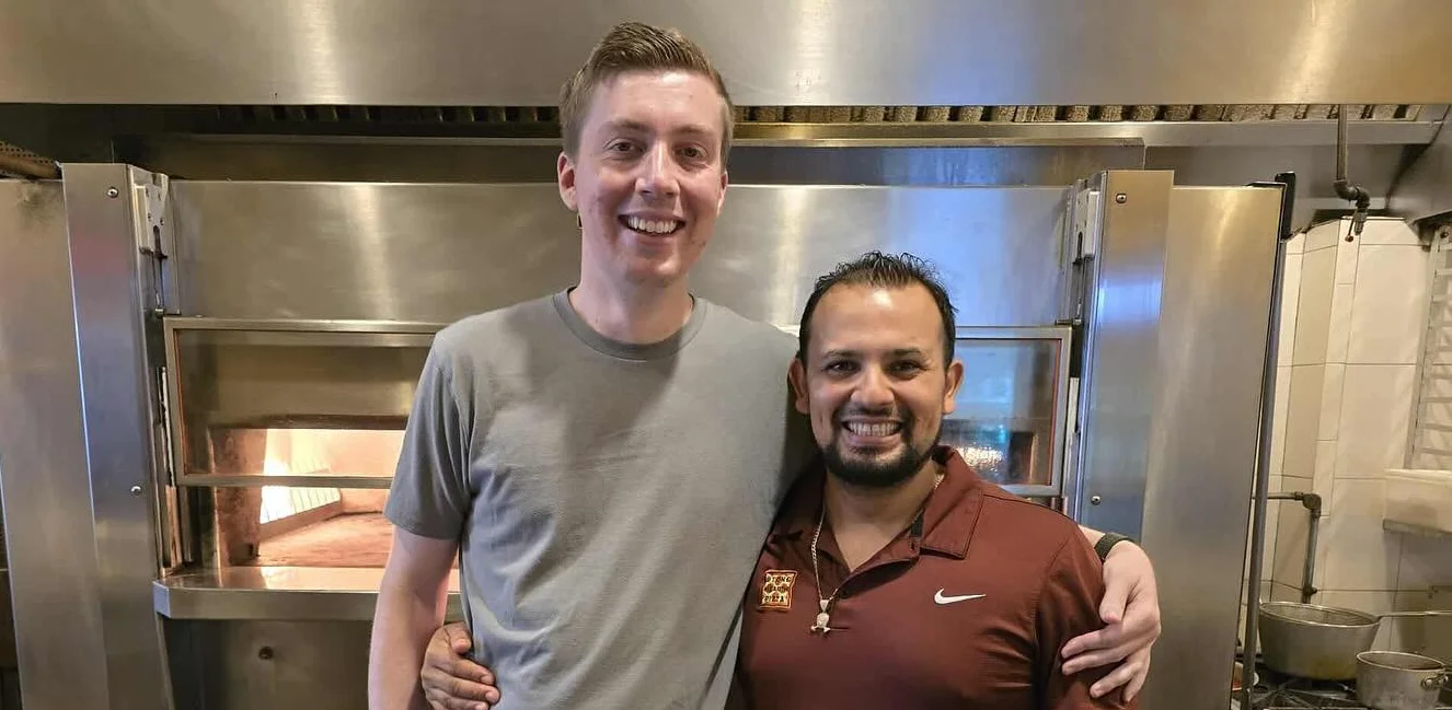 Two chefs smiling in a restaurant kitchen with a wood-fired oven