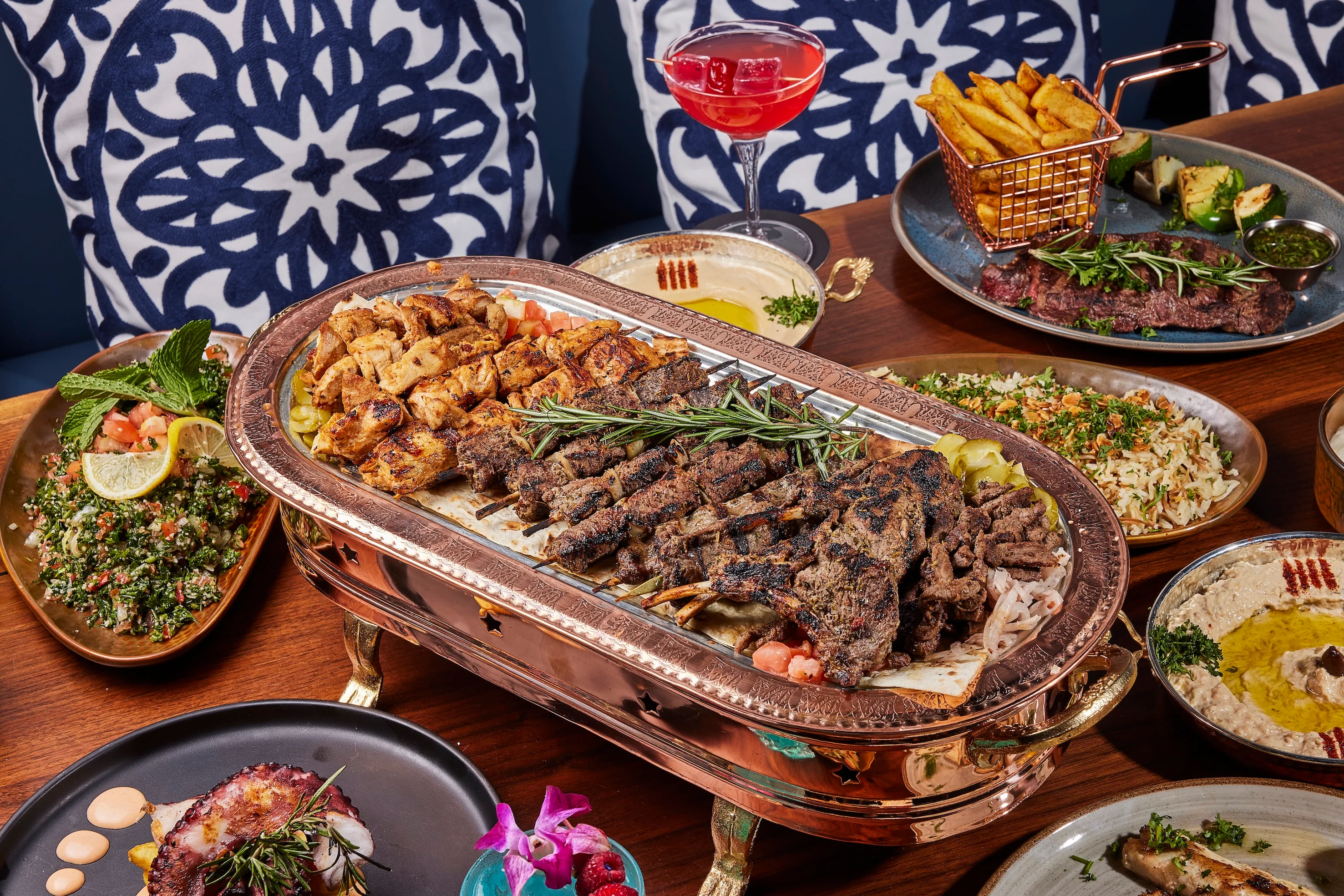 A vibrant array of Mediterranean dishes on a wooden table, featuring grilled meats on a large platter, colorful drinks, hummus, and dips, with decorative blue-patterned cushions in the background.