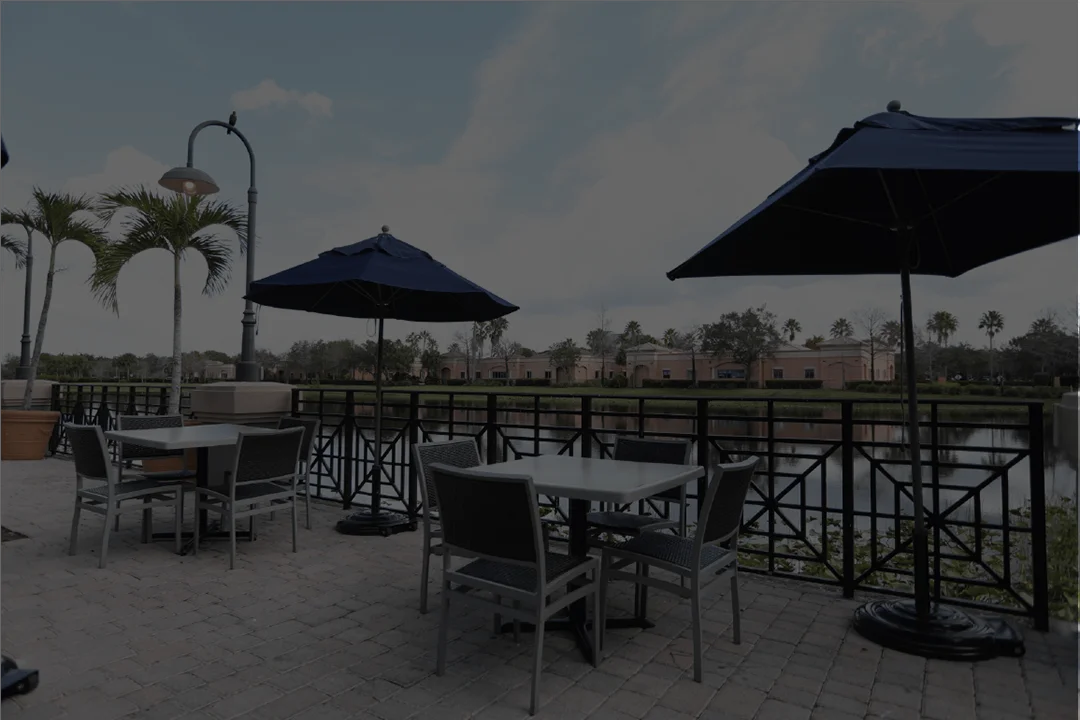 Wide-angle view of an outdoor restaurant patio with grey tables and black chairs under navy blue umbrellas, overlooking a calm waterfront with palm trees and Mediterranean-style buildings.