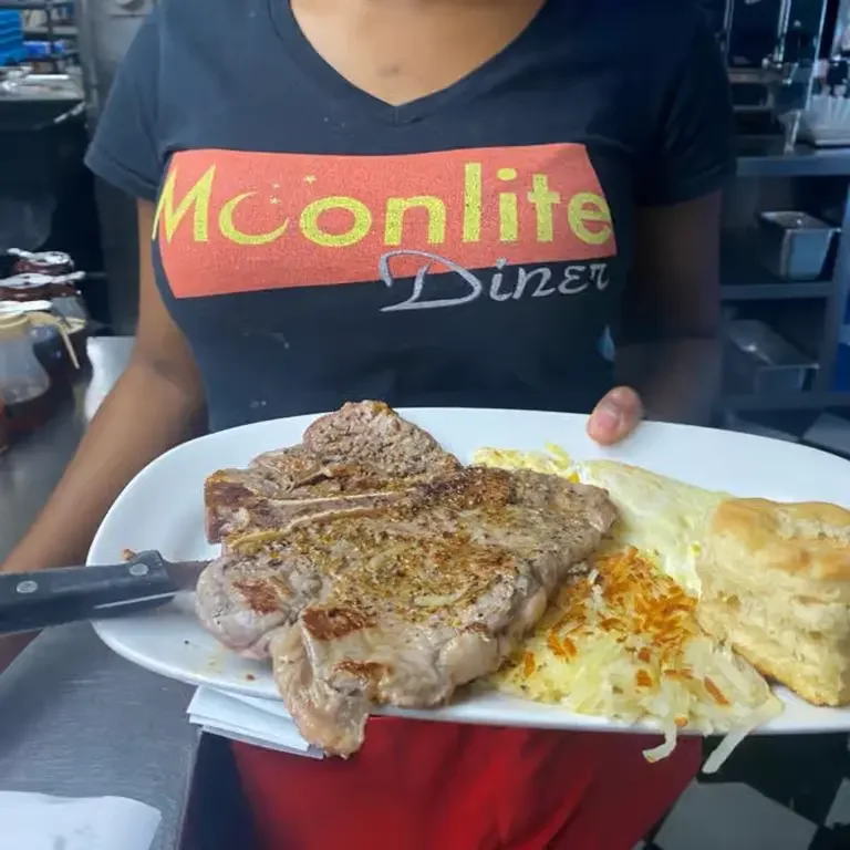 Moonlite Diner server presenting a grilled pork chop breakfast with eggs, crispy hash browns, and a freshly baked biscuit.