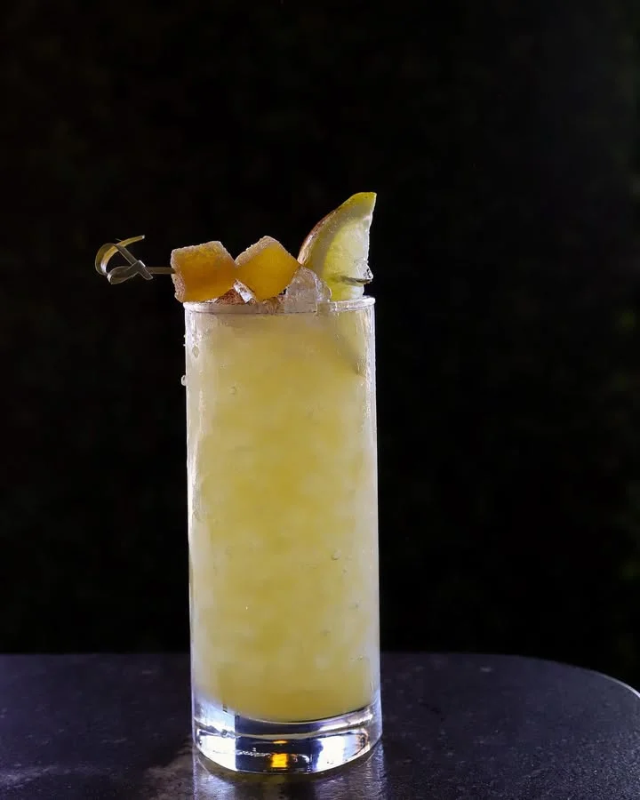 A tall glass of cocktail with a yellow hue, garnished with lemon and orange slices on top.