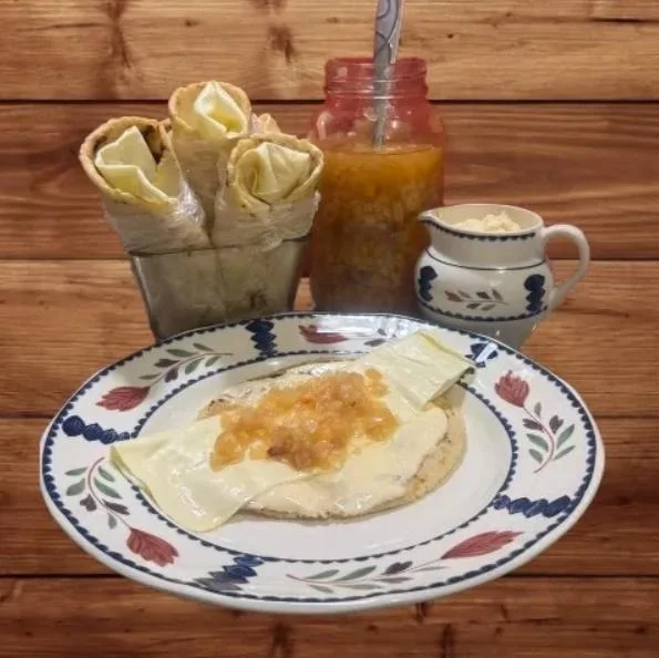 A plate with a tortilla topped with cheese and a sweet fruit topping, accompanied by rolled pastries and a jar of fruit preserve.