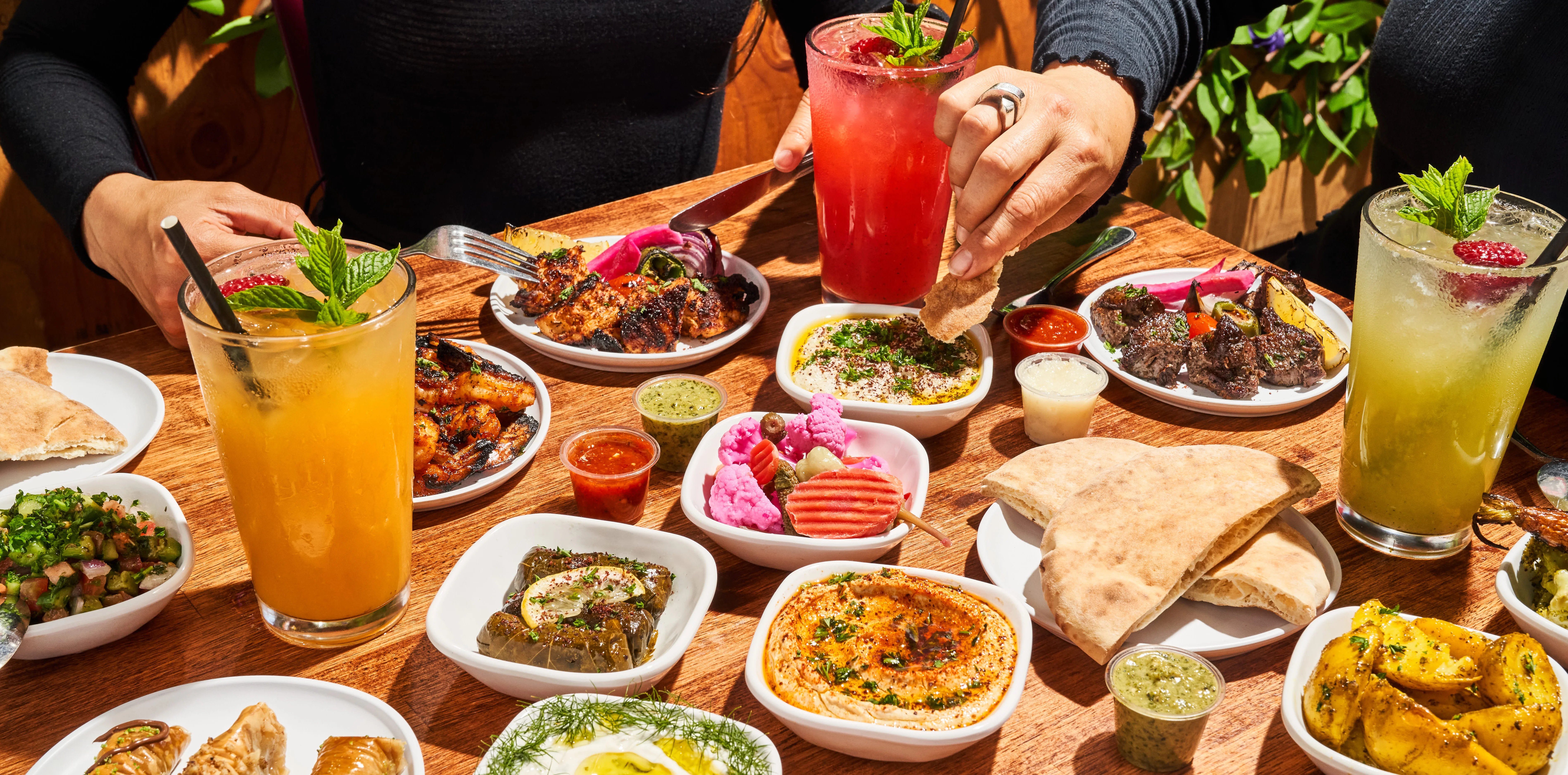 A table full of grilled meats, fresh-squeezed juices, dips, and pita