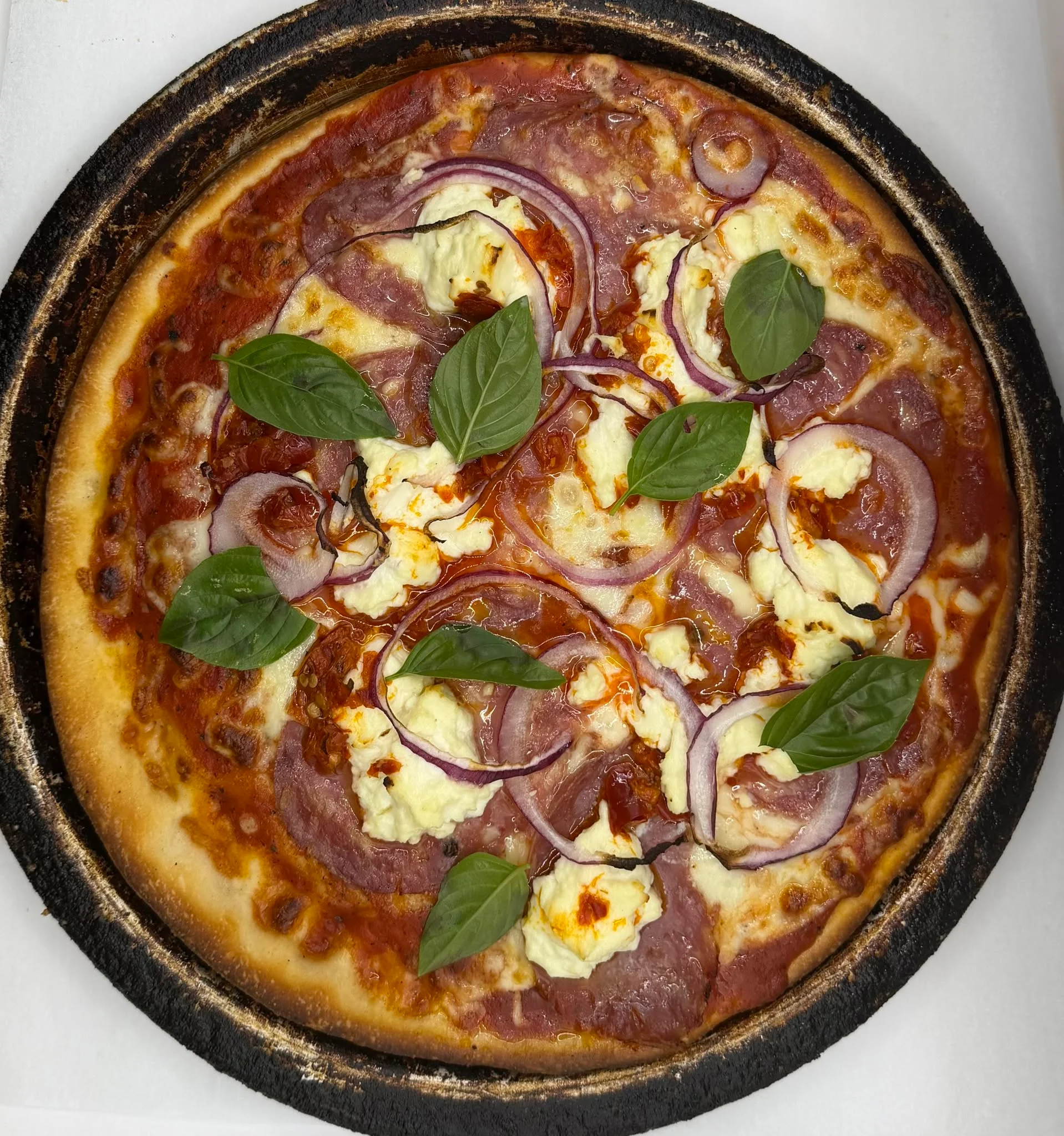 A delicious pizza topped with red onions, fresh basil, ricotta cheese, and a savory sauce, served on a dark-edged crust.