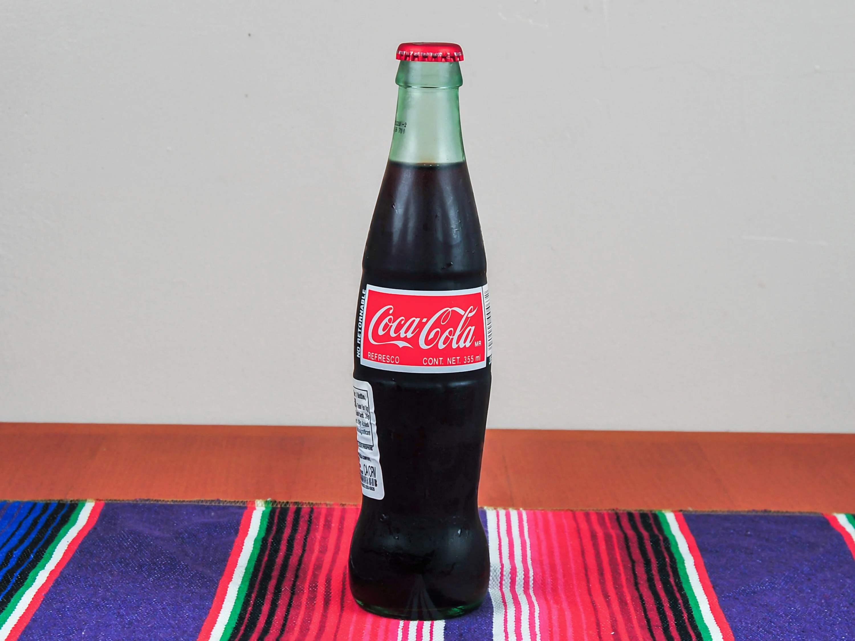 A glass bottle of Coca-Cola is displayed on a colorful tablecloth, showcasing its classic red and white label.