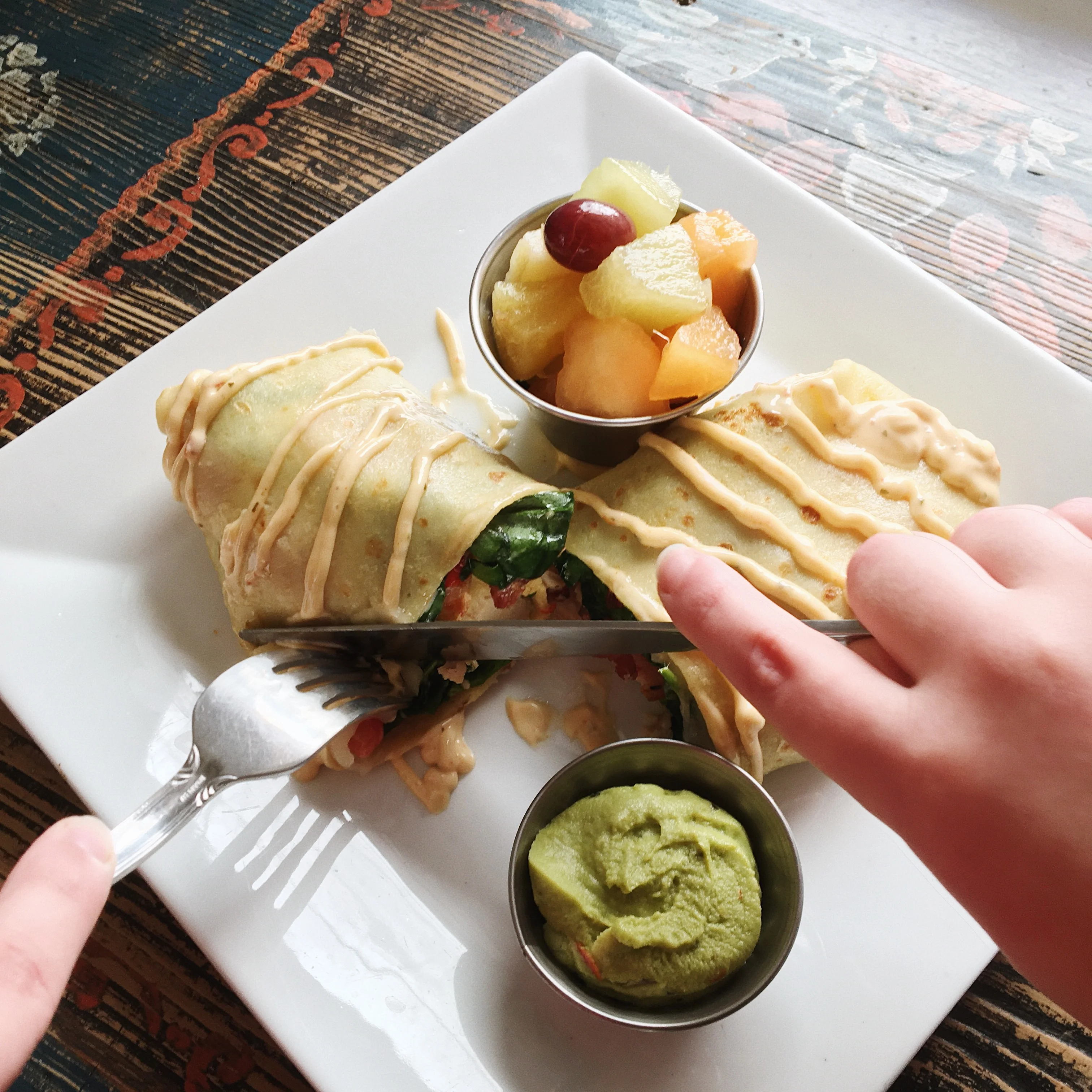 A plate of freshly made burritos drizzled with sauce, accompanied by a side of guacamole and a fruit medley served in a small bowl.