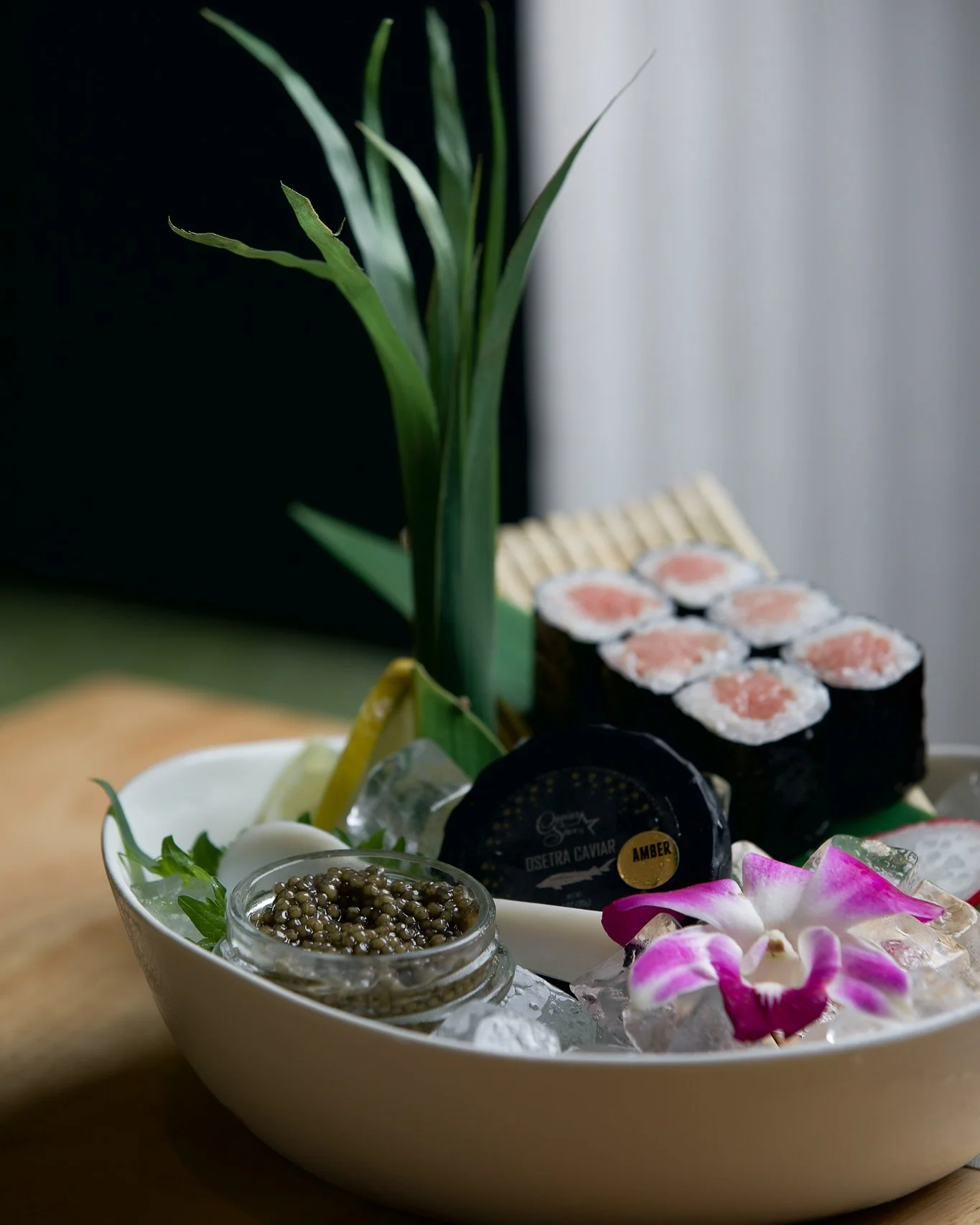 An upscale seafood presentation in a white bowl filled with ice, featuring six pieces of tuna maki sushi rolls on a bamboo mat, an open glass jar of Osetra caviar alongside its black lid, tall green leaves, and a vibrant purple orchid flower garnish.