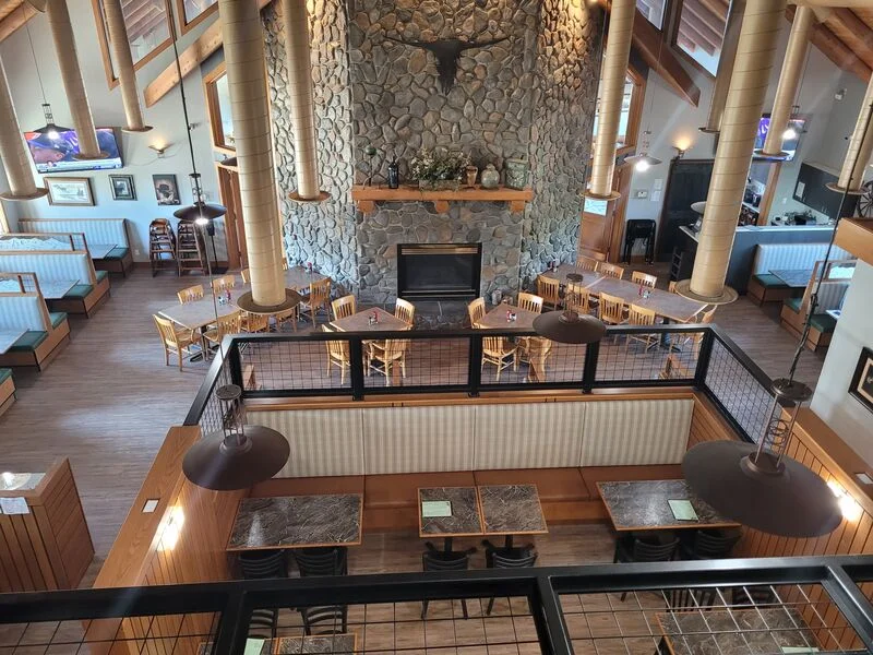 A spacious dining area featuring wooden furniture, booths, and large windows allowing natural light to fill the space. The design includes rustic elements like stone and wood.