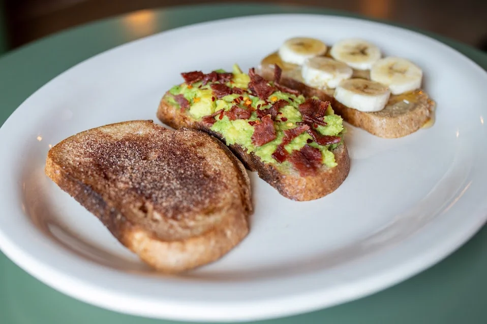 Three slices of gourmet toast. One is topped with avocado and bacon bits, another features sliced bananas, and the last has a sweet topping, all arranged on a white plate.
