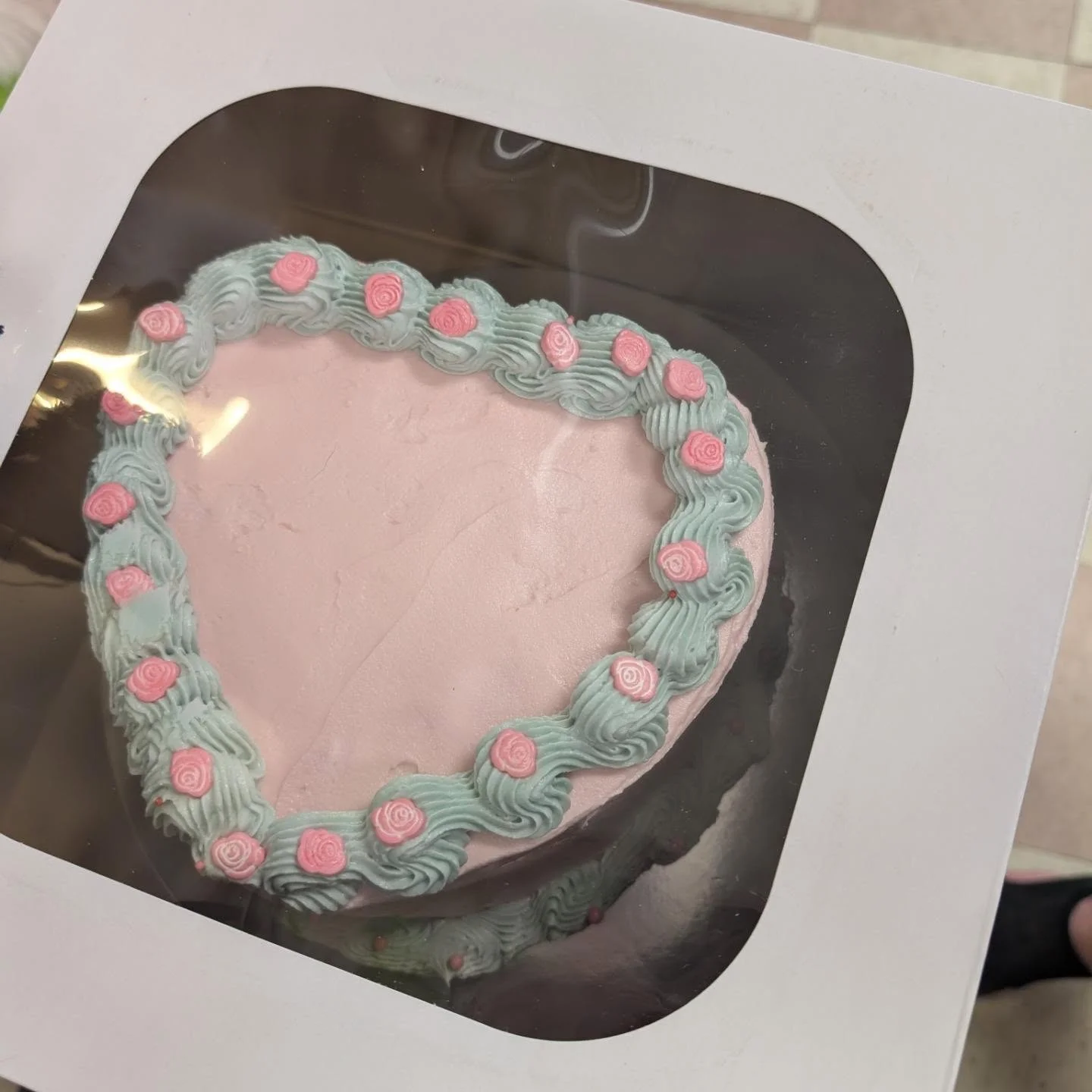 A heart-shaped cake beautifully decorated with pink and blue frosting, presented in a takeout box.
