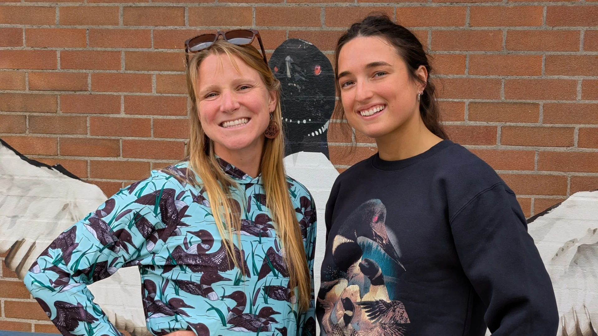 Two smiling women posing against a colorful brick wall mural