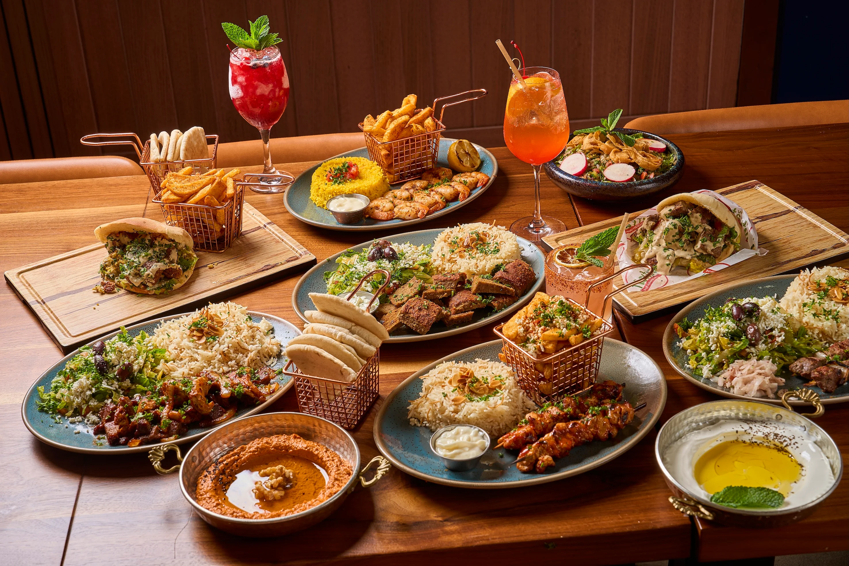 A colorful spread of Middle Eastern dishes on a wooden table, featuring kebabs, rice, salads, pita, vibrant cocktails, and dipping sauces. Cozy, inviting ambiance.