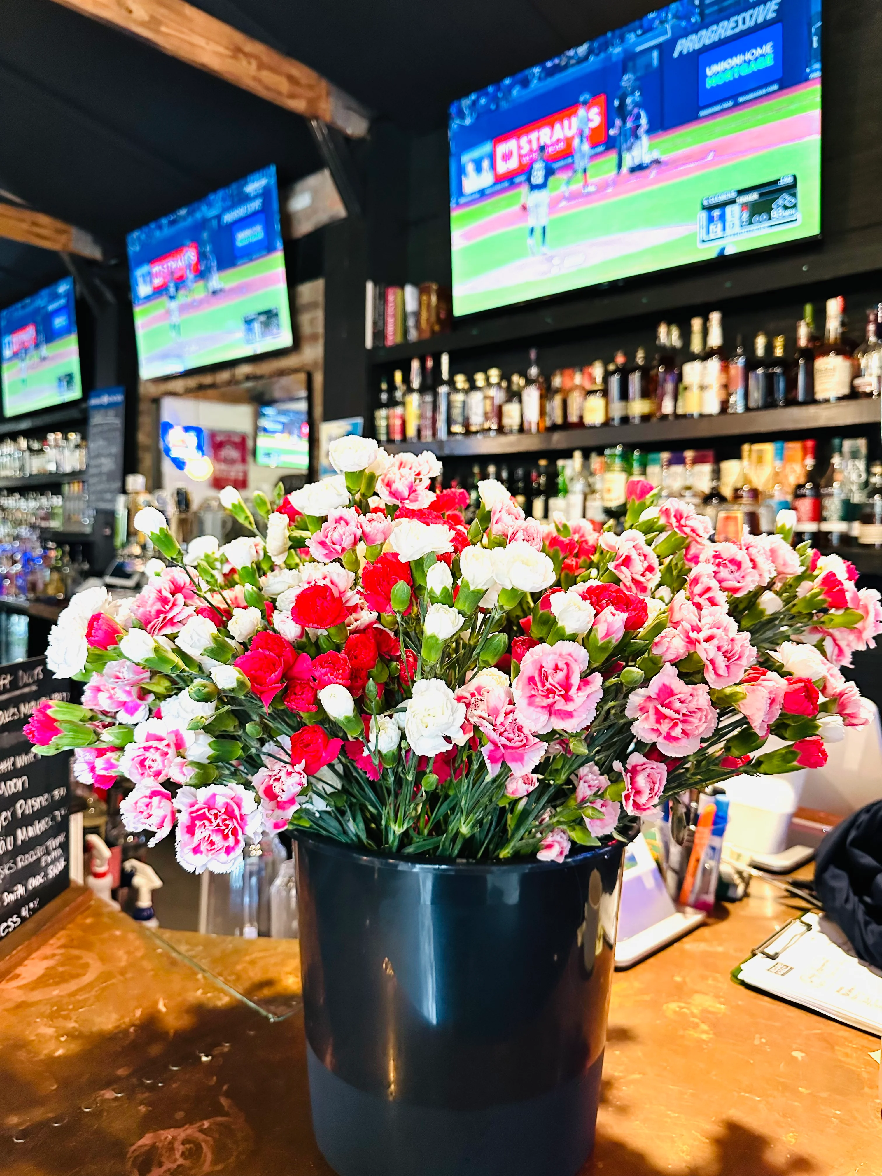 A floral arrangement of pink and red carnations in a black vase, set against a backdrop of bar shelves and televisions showing a sports game.