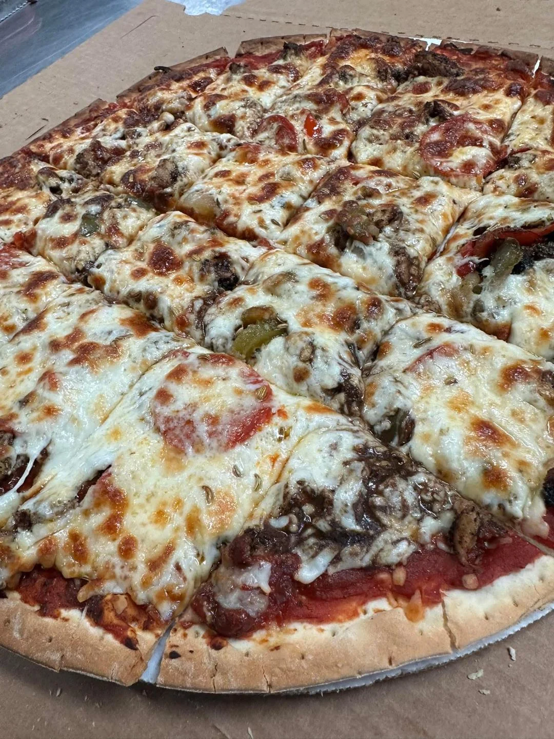 A pizza with melted cheese and various toppings including meat and vegetables, cut into slices and presented in a cardboard box.