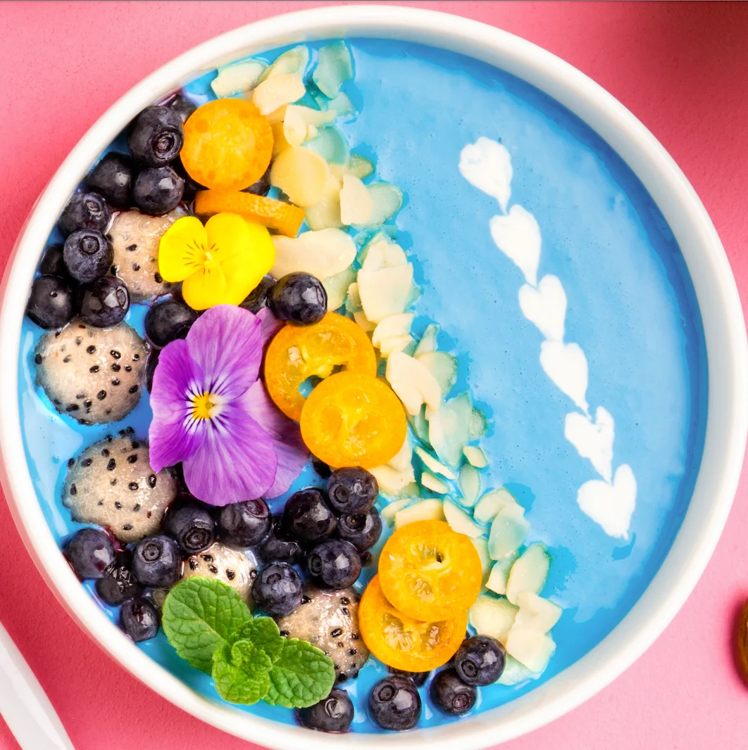 A vibrant blue smoothie bowl topped with an assortment of fruits including blueberries, kumquats, and almonds, along with edible flowers for decoration.