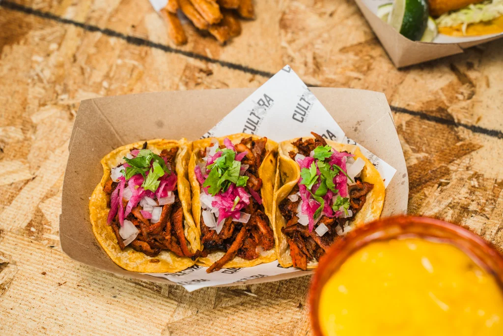 Three tacos topped with onions, cilantro, and pink pickled onions next to a drink.