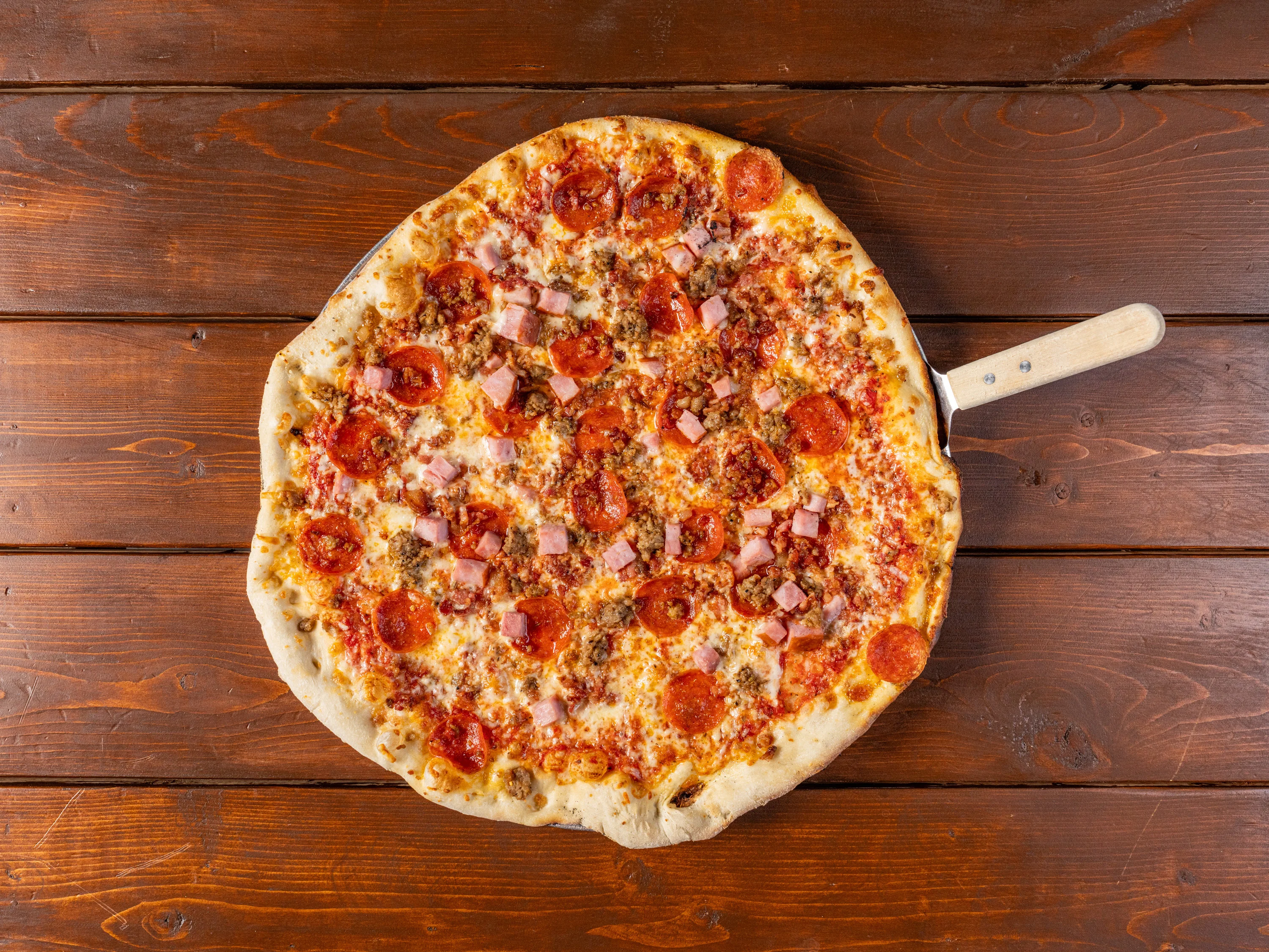 An appetizing Butcher Shop Pizza loaded with a generous topping of pepperoni, Italian sausage, ham, and bacon, showcasing a golden crust and a melty cheese blend, served on a rustic wooden table indicative of the hearty and savory flavors awaiting.