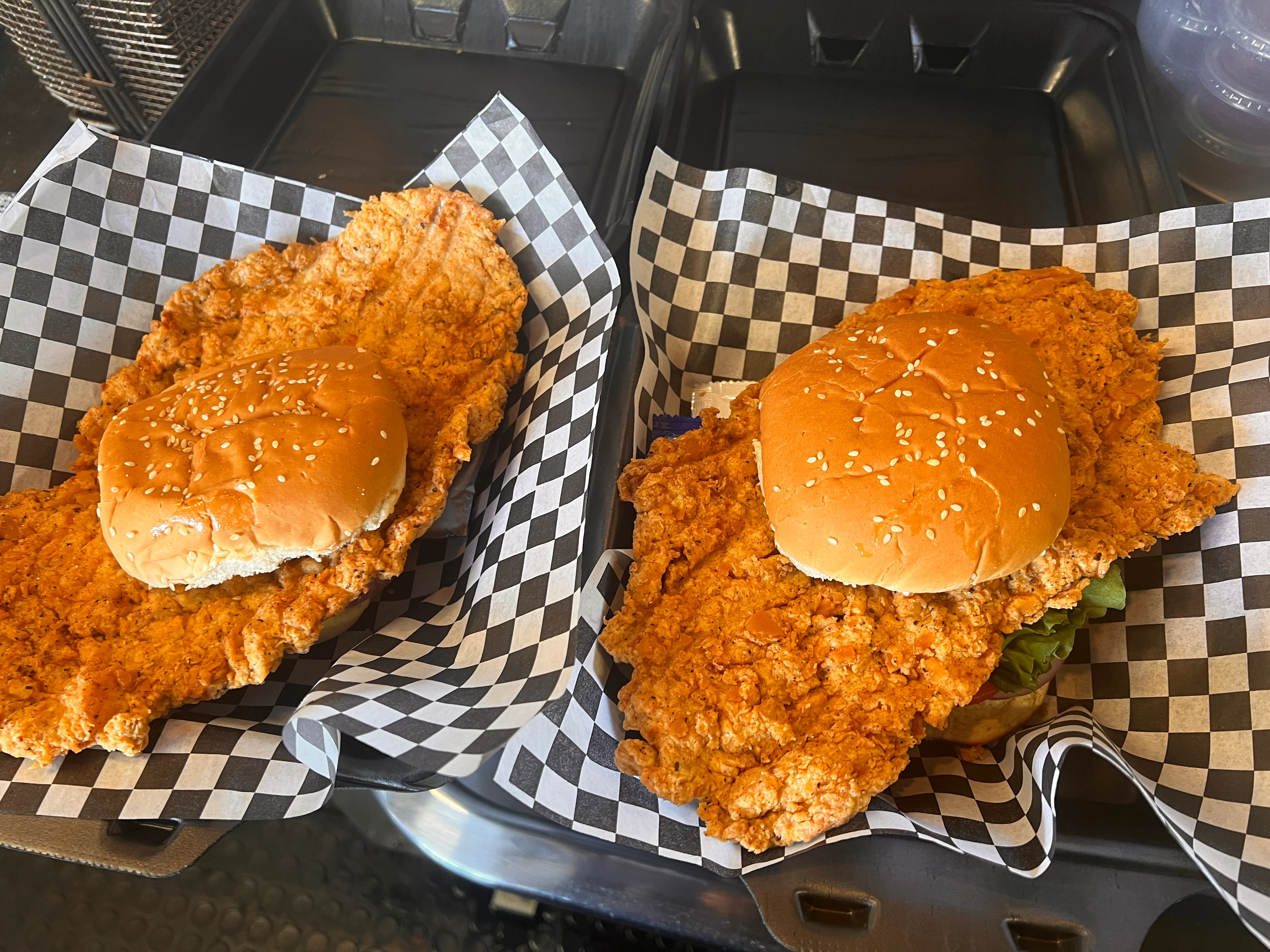Two large fried chicken sandwiches with sesame seed buns, served in checkered paper.