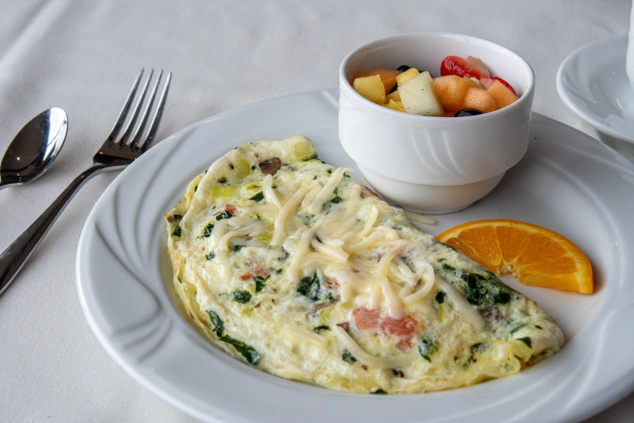 A delicious omelette filled with spinach, tomatoes, and cheese, served alongside a bowl of mixed fruit and a slice of orange.