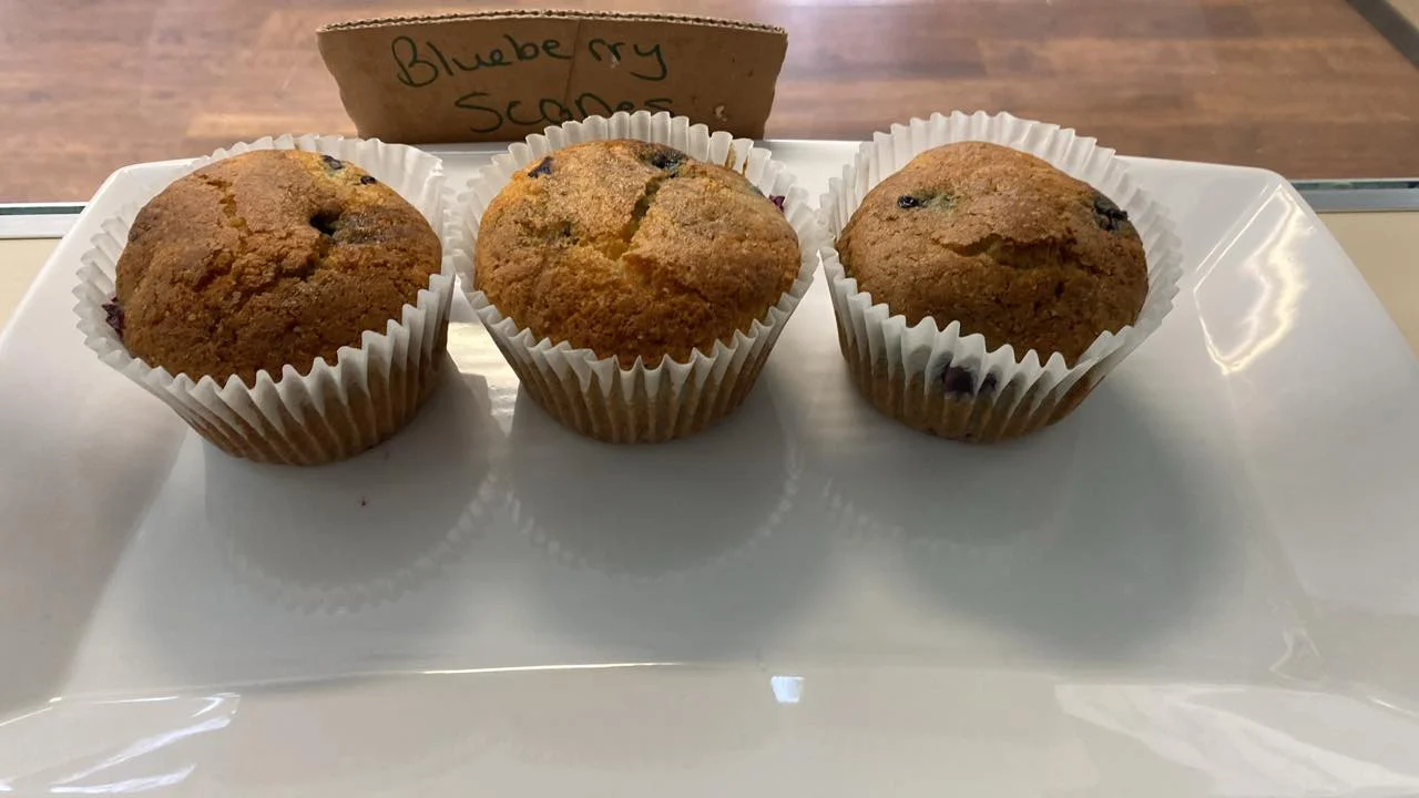 Three blueberry scone muffins in paper liners, presented on a white plate. They are golden and have blueberries peeking through.