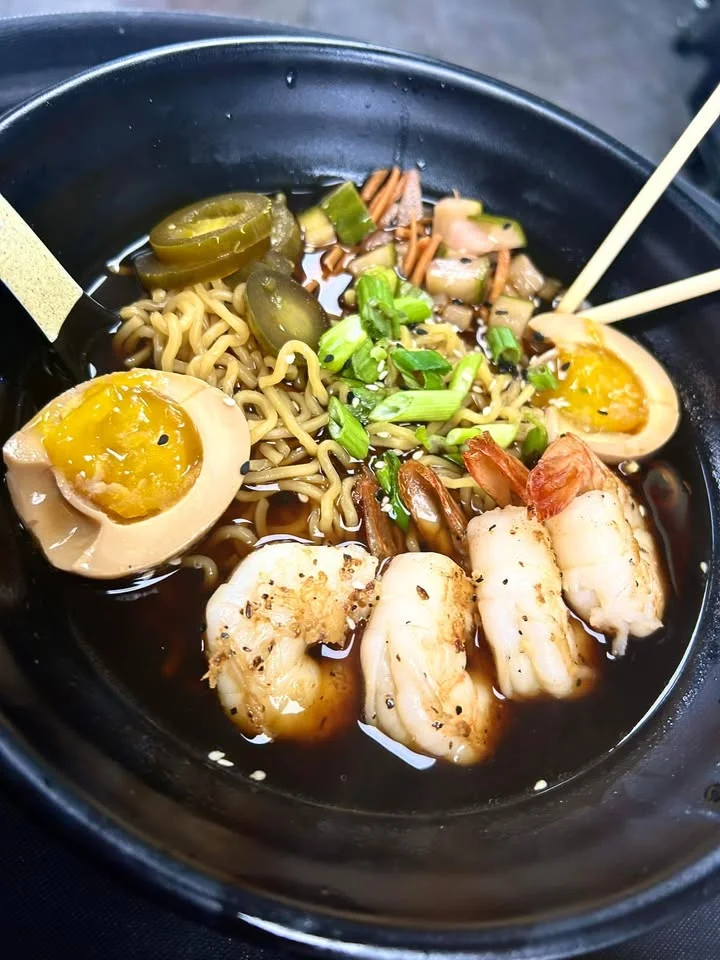 A bowl of ramen featuring noodles, shrimp, soft-boiled egg, green onions, and pickled vegetables in a rich broth.