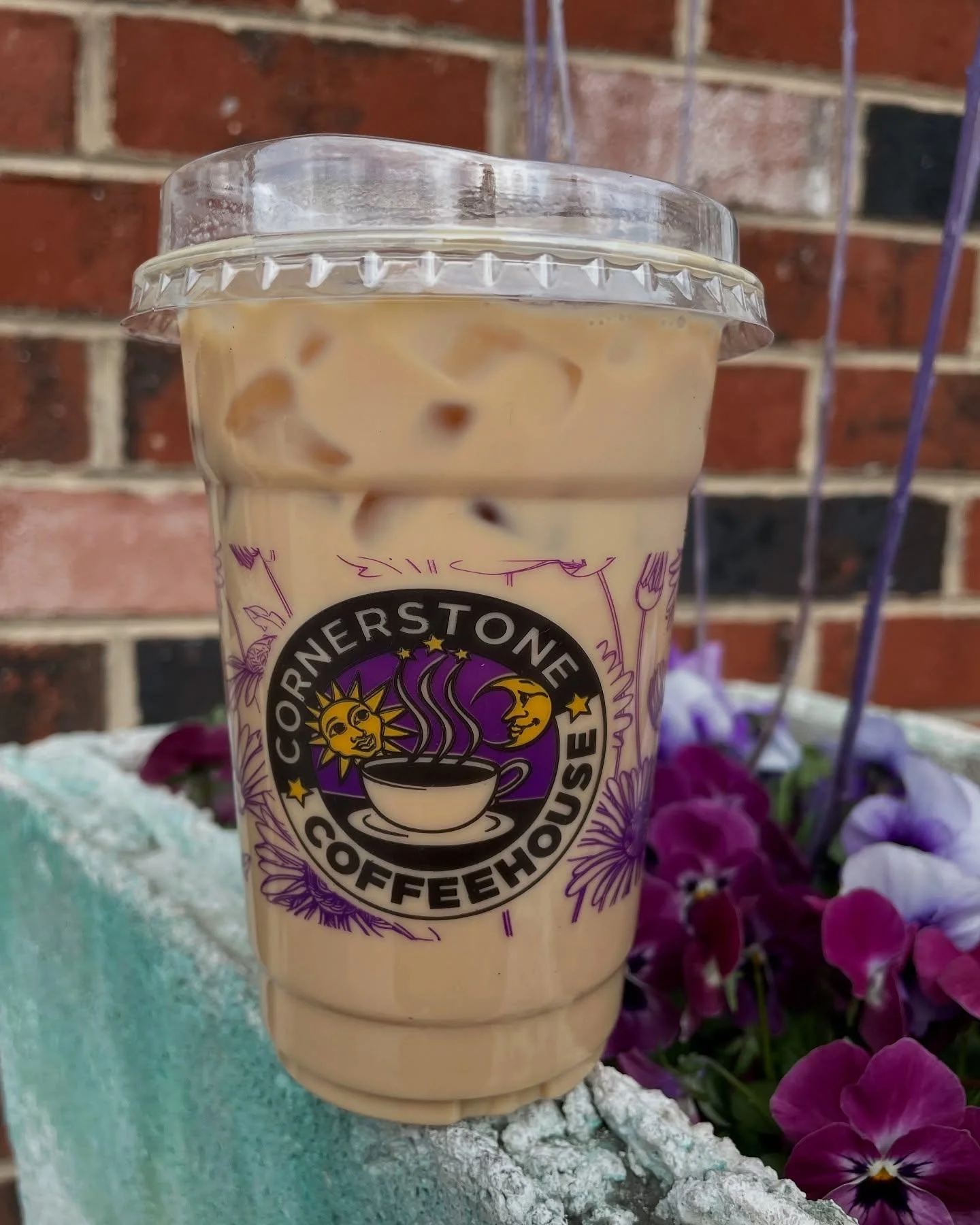 A clear plastic cup filled with iced coffee, featuring a logo and sitting near purple flowers.