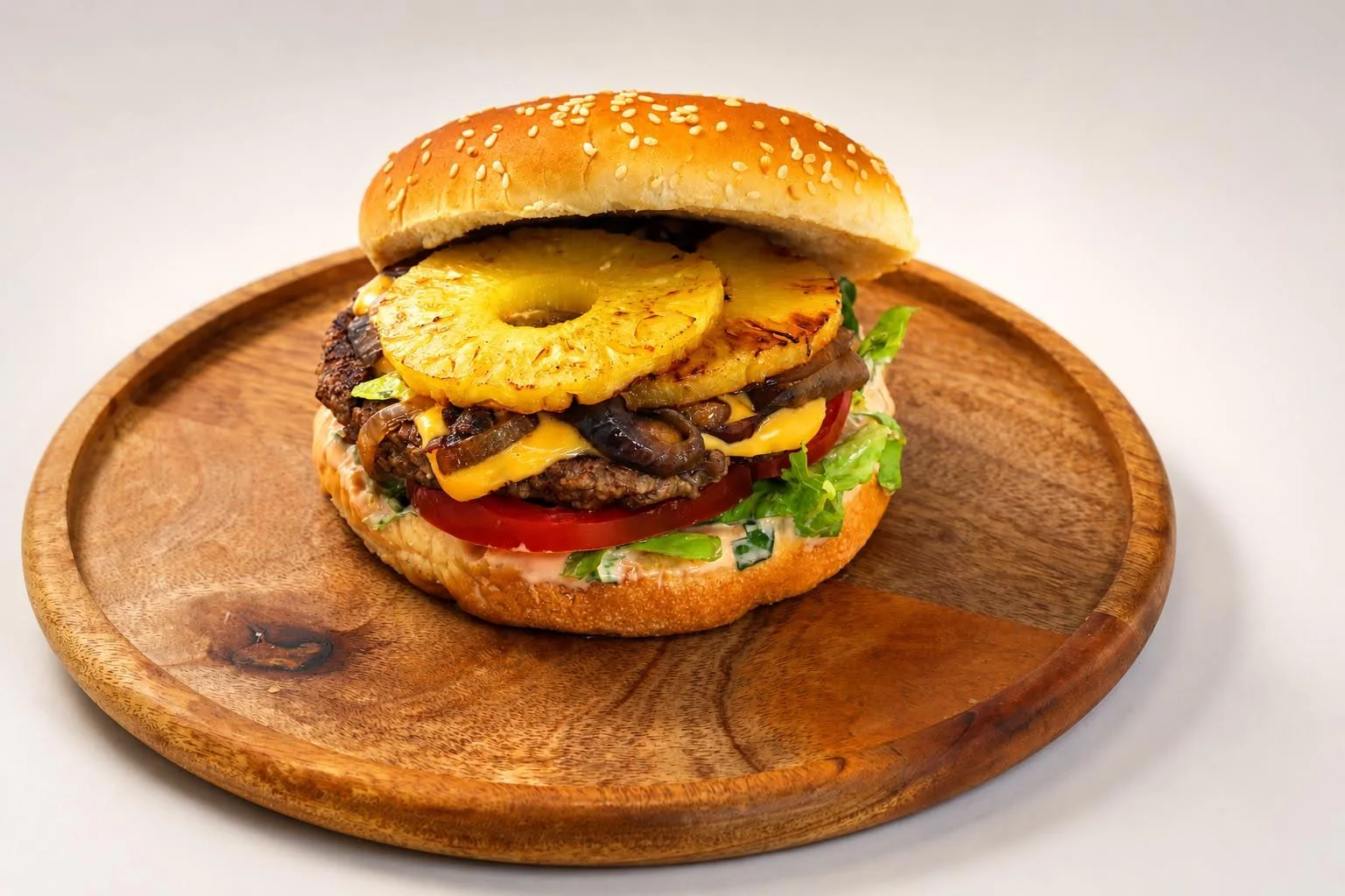 A hearty burger stacked with a beef patty, cheddar cheese, grilled pineapple, lettuce, and tomato, served on a sesame seed bun on a wooden plate.