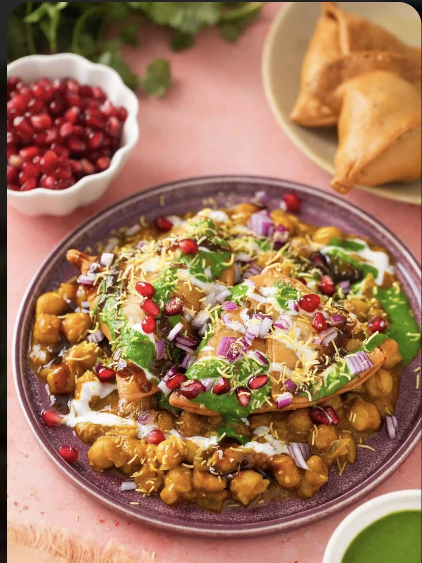 A vibrant plate of chaat with crispy components, garnished with yogurt, green chutney, and pomegranate seeds, alongside samosas and a bowl of pomegranate.