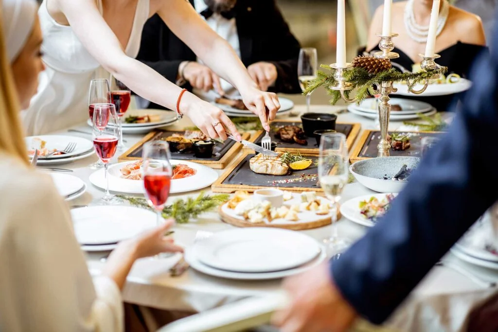 A elegantly set dining table featuring a variety of dishes, glasses of red and sparkling beverages, and candlelight for ambiance. Guests are engaged in serving food from the central platter.