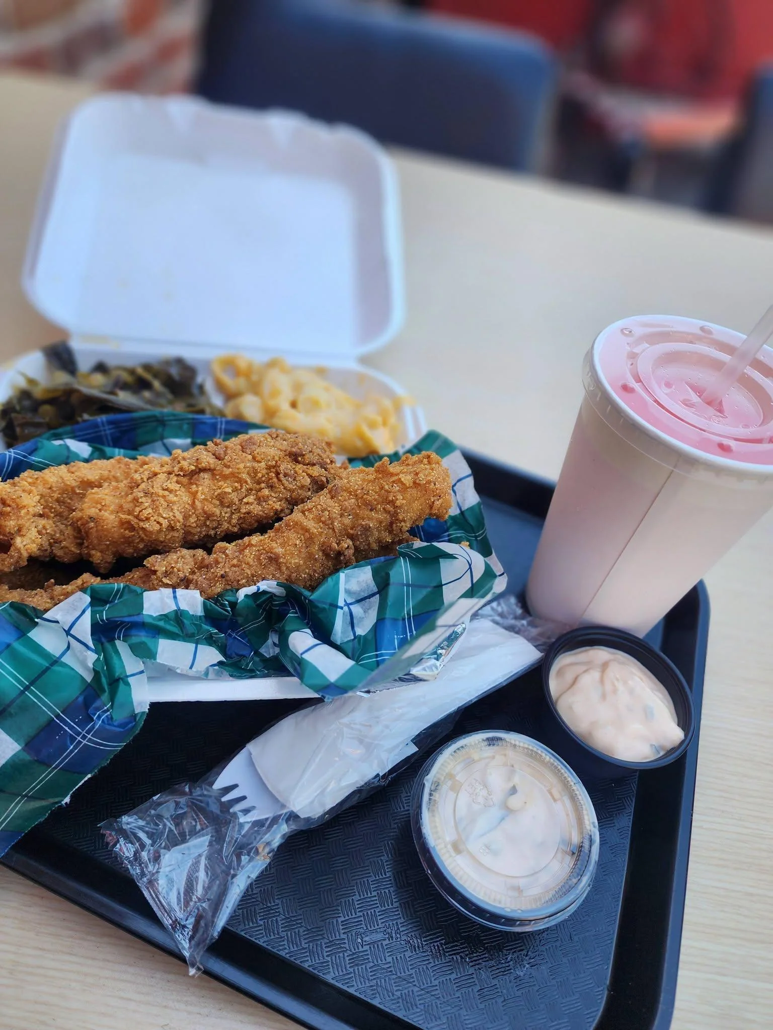 A meal composed of crispy fried chicken tenders, sides of greens and macaroni, and a strawberry milkshake presented on a tray.