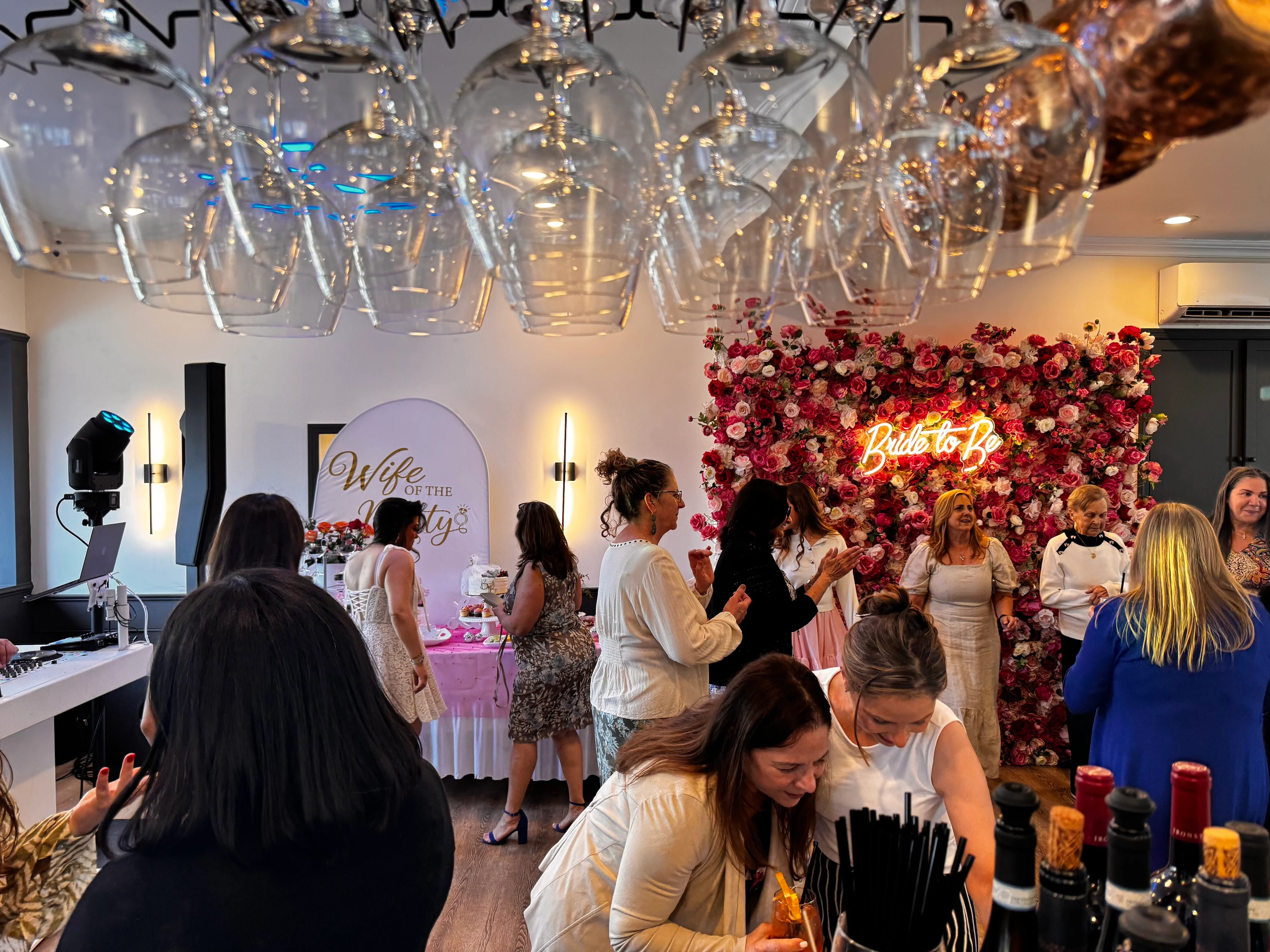 A lively bridal shower with elegantly dressed women socializing. A neon "Bride to Be" sign is set against a red floral wall, creating a festive atmosphere.