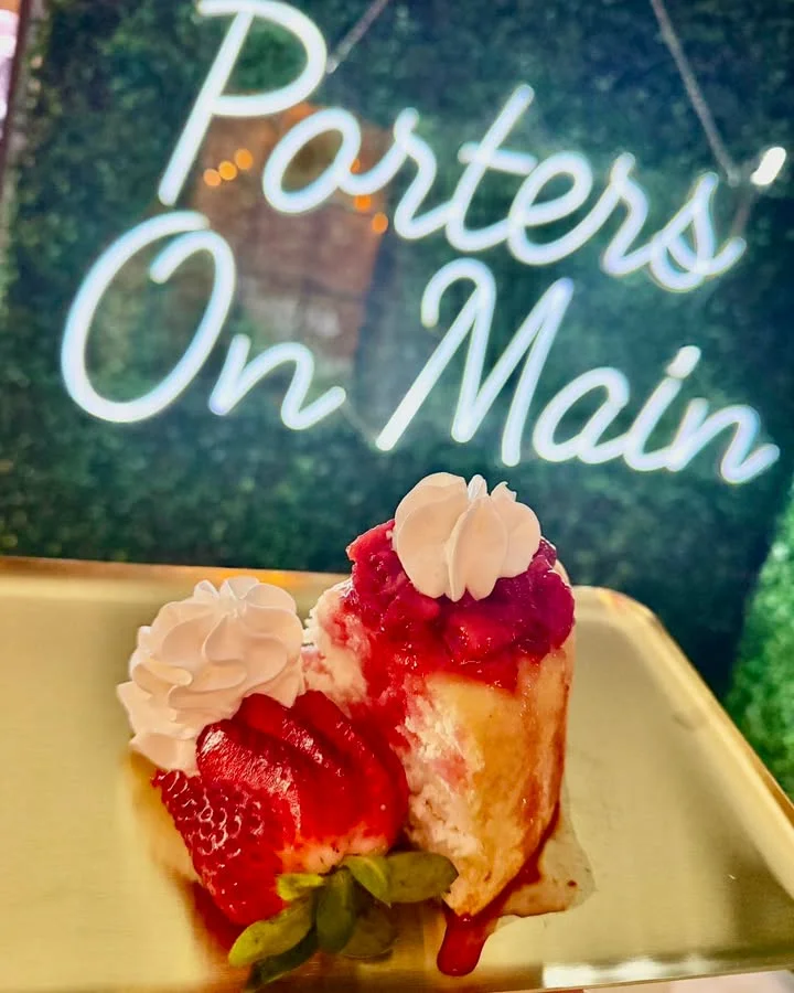 A slice of strawberry cheesecake topped with whipped cream and a fresh strawberry, placed on a gold plate in front of a sign.