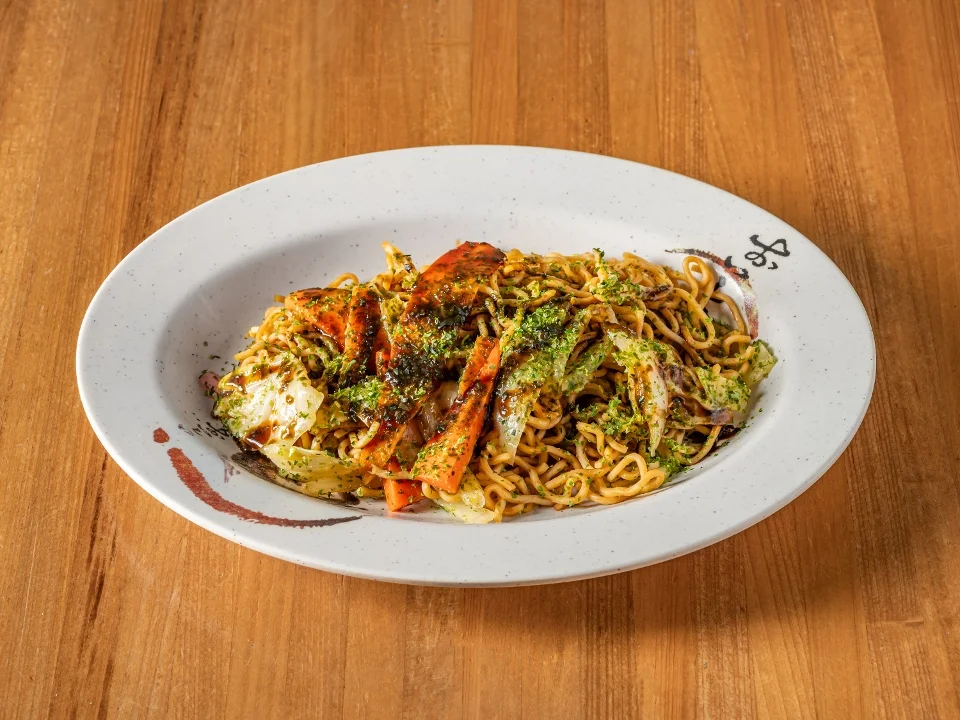 A plate of stir-fried noodles topped with grilled vegetables and a sprinkle of herbs. The dish features colorful, sautéed carrots and cabbage.