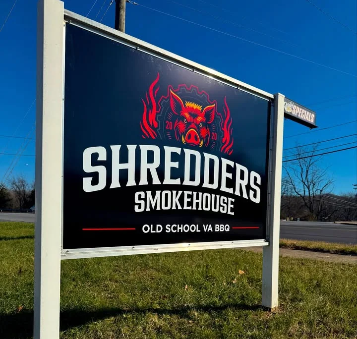 A roadside sign displaying the smokehouse name and logo, situated in a green lawn under a clear blue sky.