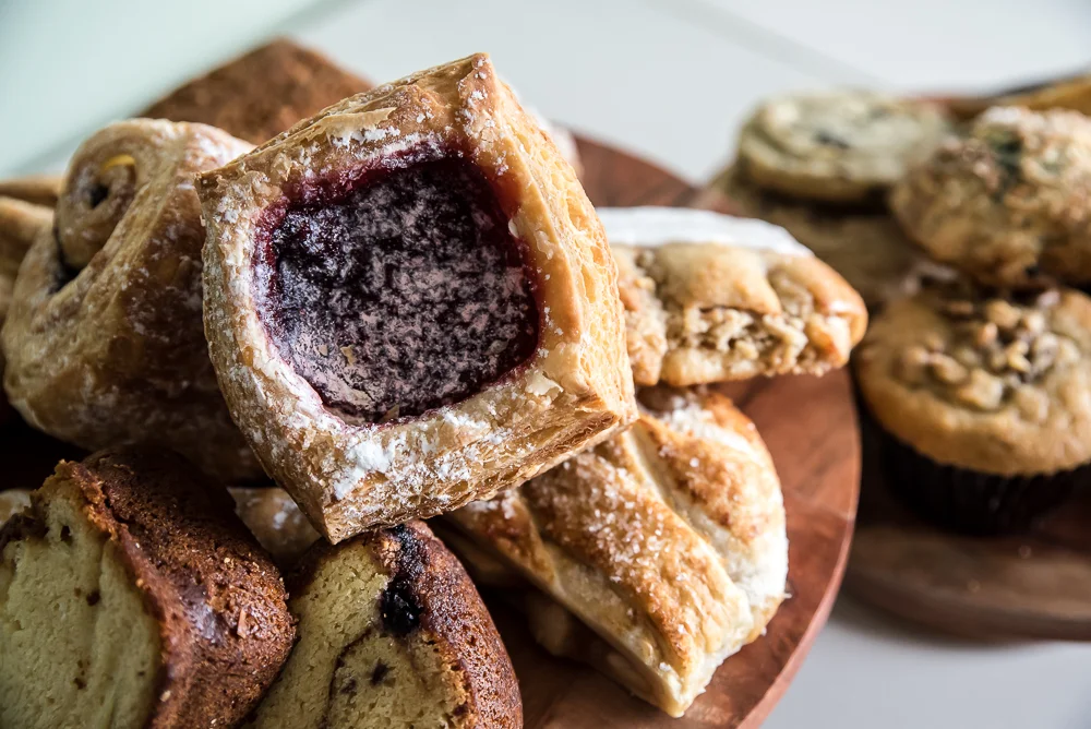 A platter of assorted pastries, including a square pastry filled with berry jam, muffins, and a slice of cake.