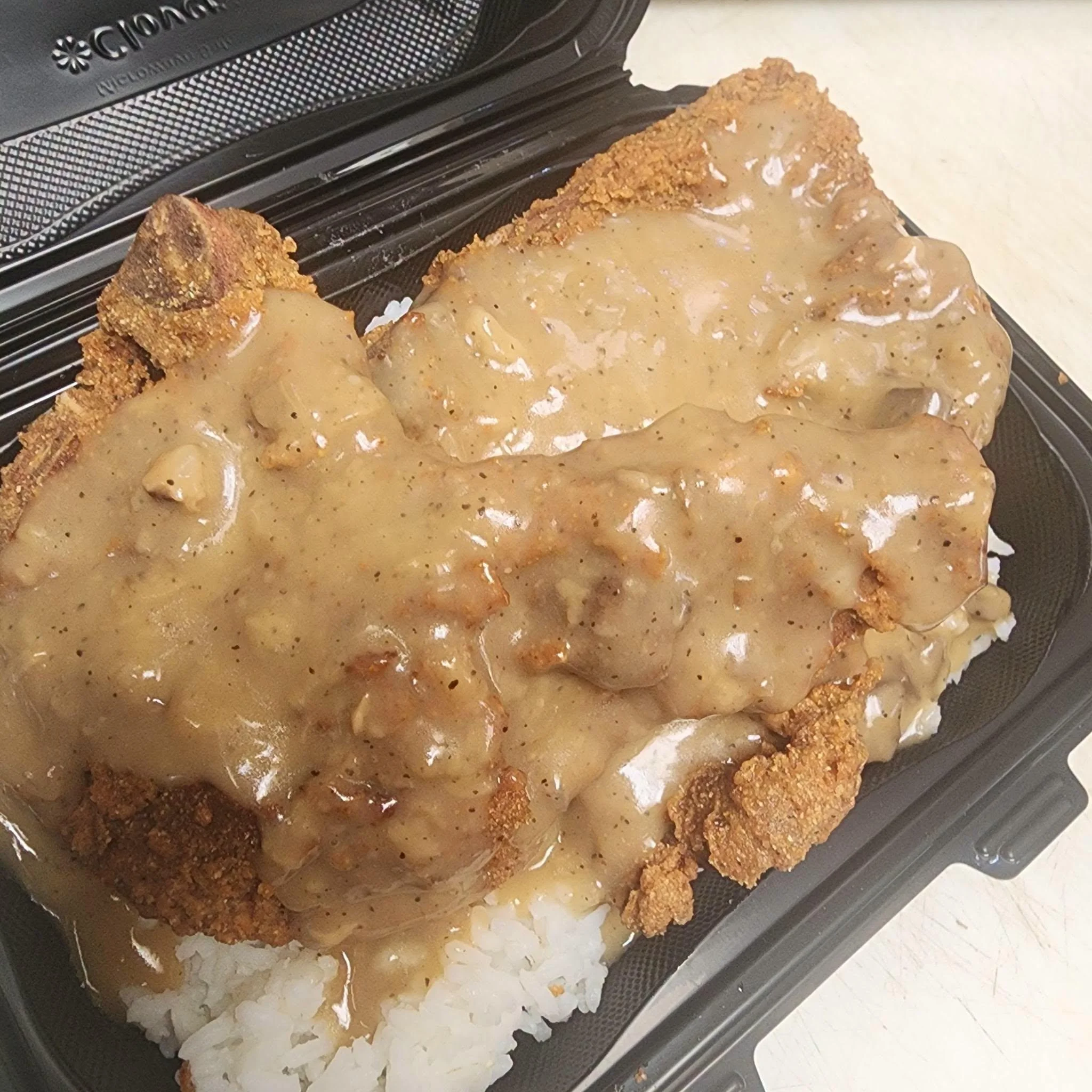 A serving of fried chicken topped with gravy, served over a bed of white rice in a takeout container.