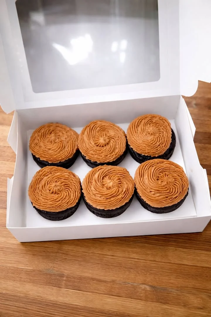 A box containing six chocolate cupcakes topped with a smooth, brown frosting in a spiral pattern.
