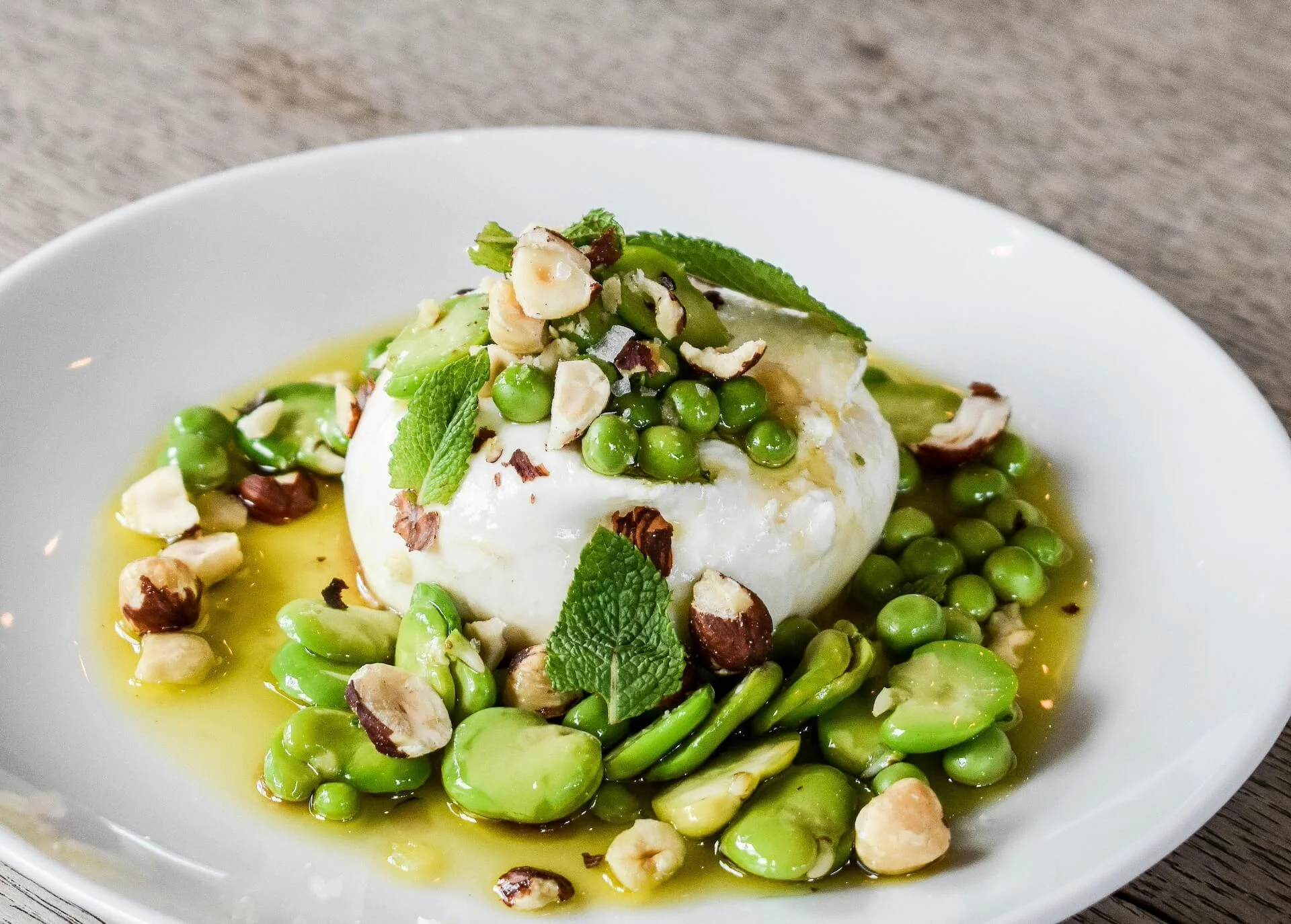 Delicious burrata cheese topped with fresh green peas, fava beans, and crunchy nuts, drizzled with olive oil and mint leaves served at Toscana on Main